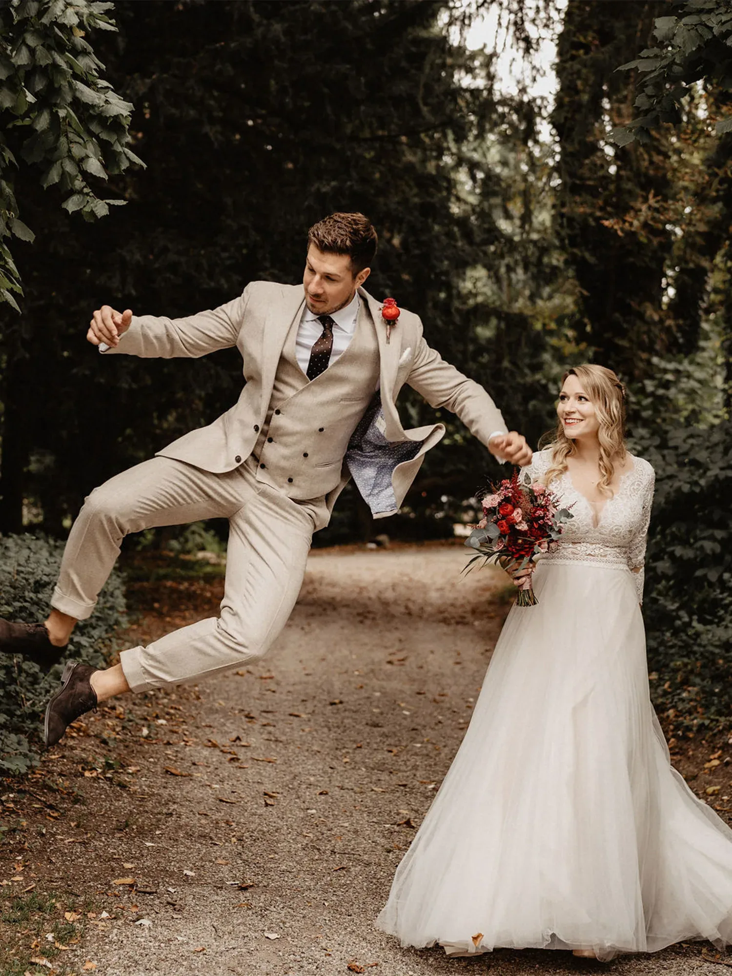 Der Bräutigam springt in die Luft, während die Braut ihn lächelnd an der Hand hält. Sie trägt ein elegantes Spitzenkleid und einen Blumenstrauß, und der Bräutigam ist in einen beigen Anzug gekleidet. Beide stehen auf einem Waldweg.