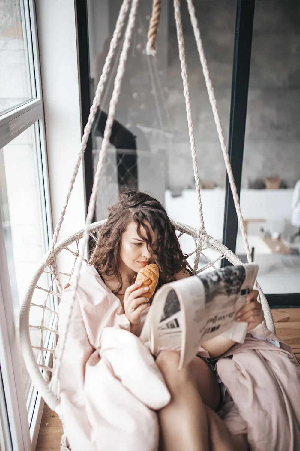 Eine Frau sitzt eingehüllt in eine Decke auf einer Hängeschaukel, isst ein Croissant und liest eine Zeitung. Sie genießt einen ruhigen Moment neben einem großen Fenster, das viel Tageslicht in den Raum lässt.