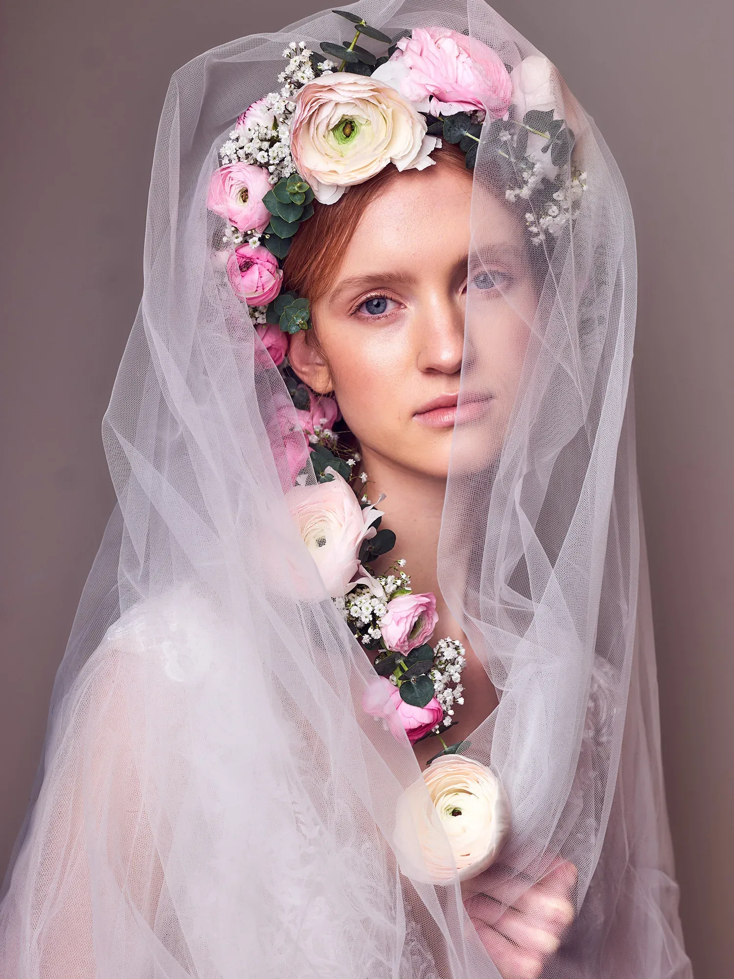 Eine Braut mit einem floralen Kranz aus Ranunkeln und Eukalyptus trägt einen luftigen Schleier, der ihr Gesicht verhüllt. Der Kranz aus rosa, weißen und cremefarbenen Blumen verleiht der Braut einen natürlichen, aber eleganten Look, während der Schleier eine geheimnisvolle Note hinzufügt.