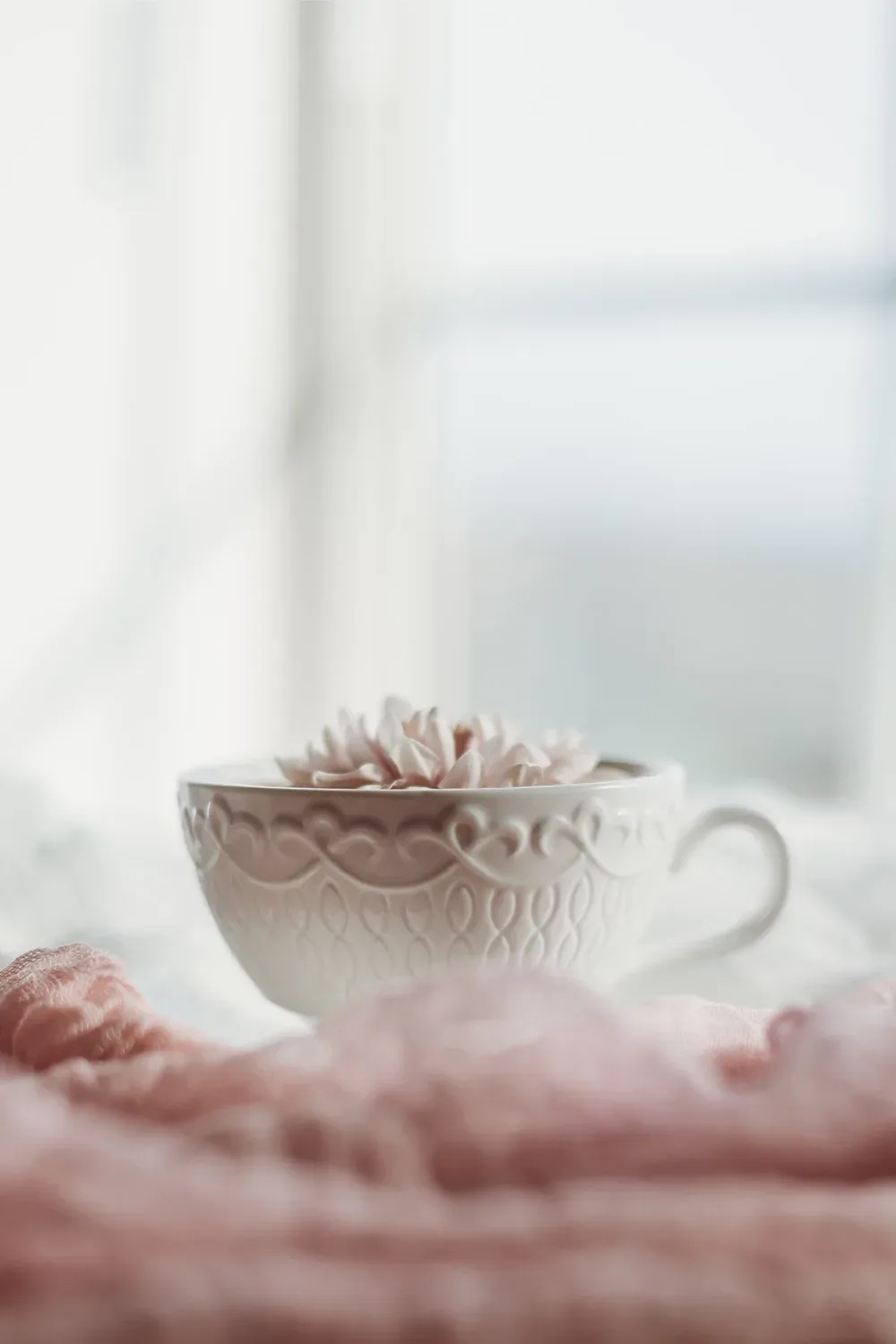 Eine dekorative, weiße Teetasse mit filigranen Mustern steht auf einem weichen, rosa Stoff. In der Tasse schwimmt eine Blüte, die eine ruhige und entspannte Atmosphäre schafft.