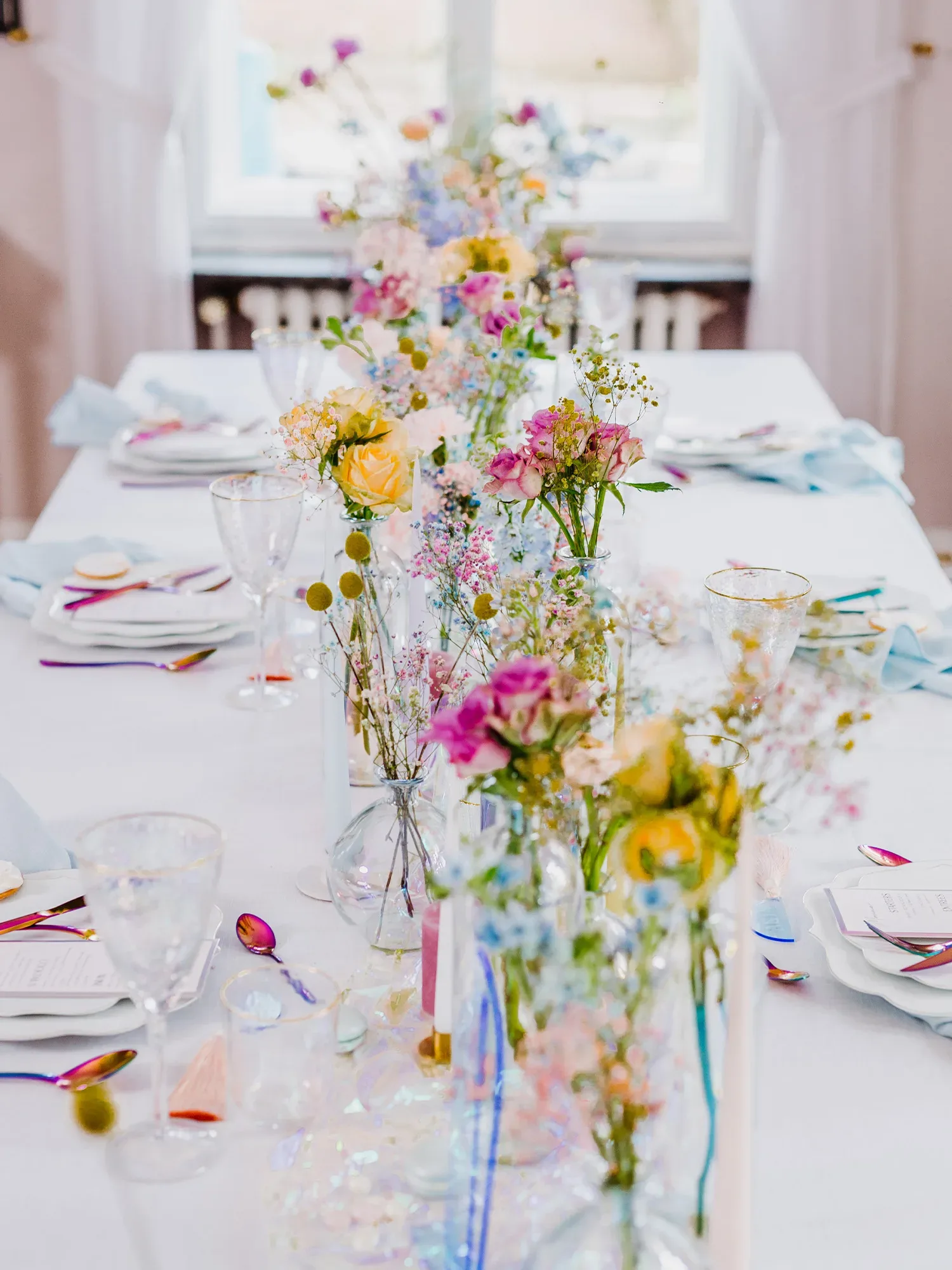 Ein langer Tisch ist elegant mit Vasen voller bunter Blumen in Pastelltönen dekoriert. Das natürliche Licht durch das Fenster im Hintergrund erhellt den Raum und sorgt für eine freundliche, festliche Atmosphäre. Die gedeckten Plätze haben funkelndes Glas und schimmerndes Besteck.
