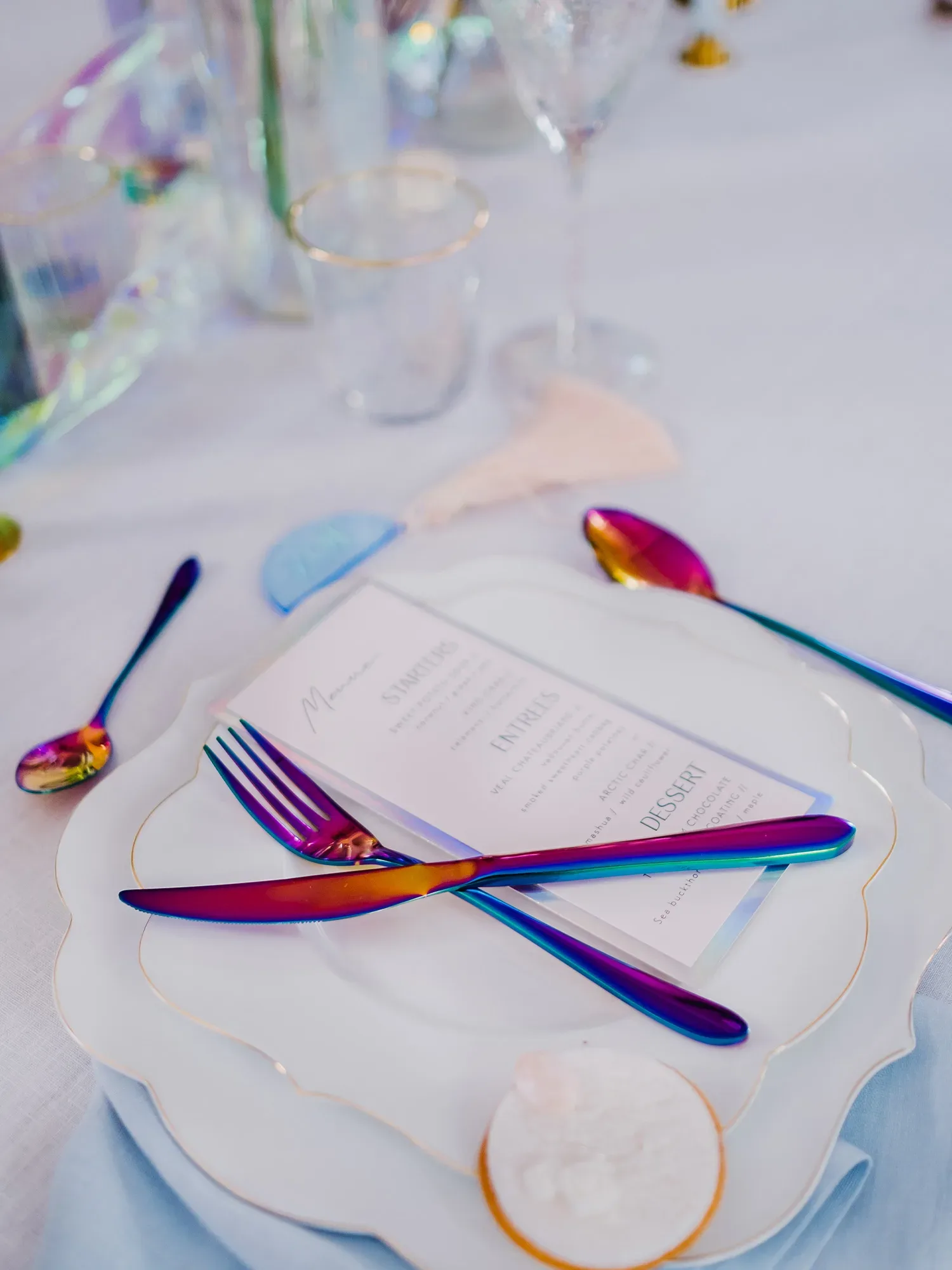 Eine Nahaufnahme eines gedeckten Tellers mit einem Menü darauf, flankiert von glänzendem Regenbogenbesteck. Die weiße Serviette und die pastellfarbenen Akzente fügen eine elegante Note hinzu. Das Glas und die Deko-Elemente schimmern leicht im Hintergrund.