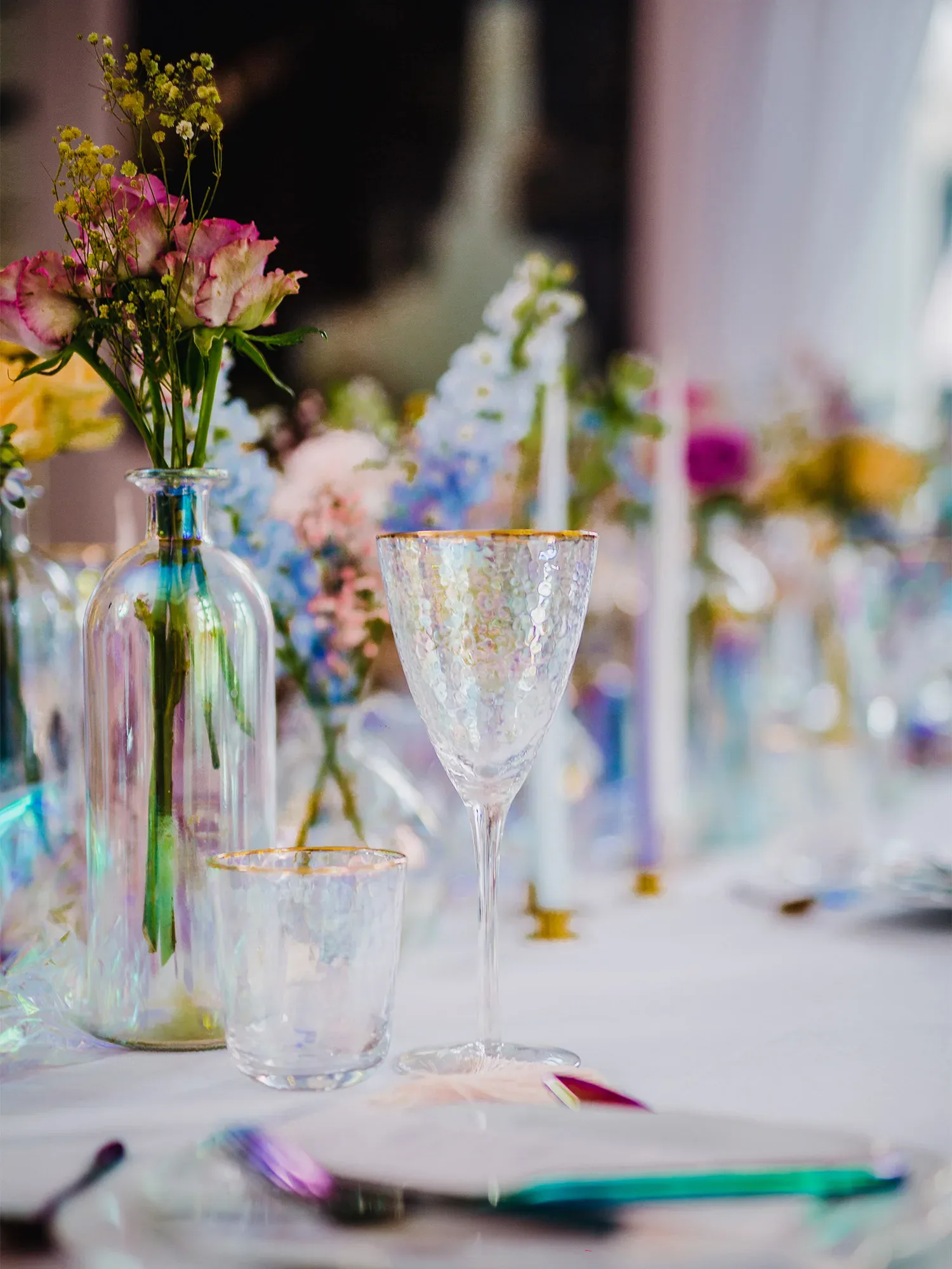 Eine Nahaufnahme eines Tisches, der mit eleganten, irisierenden Gläsern und einer Vase mit rosafarbenen Blüten in einem Glasbehälter dekoriert ist. Das goldene Dekor der Gläser verleiht der Szenerie einen Hauch von Luxus, während die farbenfrohen Blumenarrangements den Tisch lebendig wirken lassen