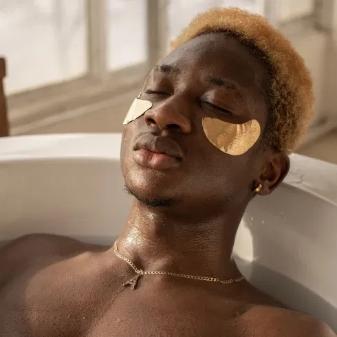 Ein Mann mit goldenen Augenpads liegt entspannt in einer mit Wasser gefüllten Badewanne. Sein Gesicht ist ruhig, seine Augen geschlossen, während weiches Tageslicht durch ein Fenster fällt und den Raum erhellt.