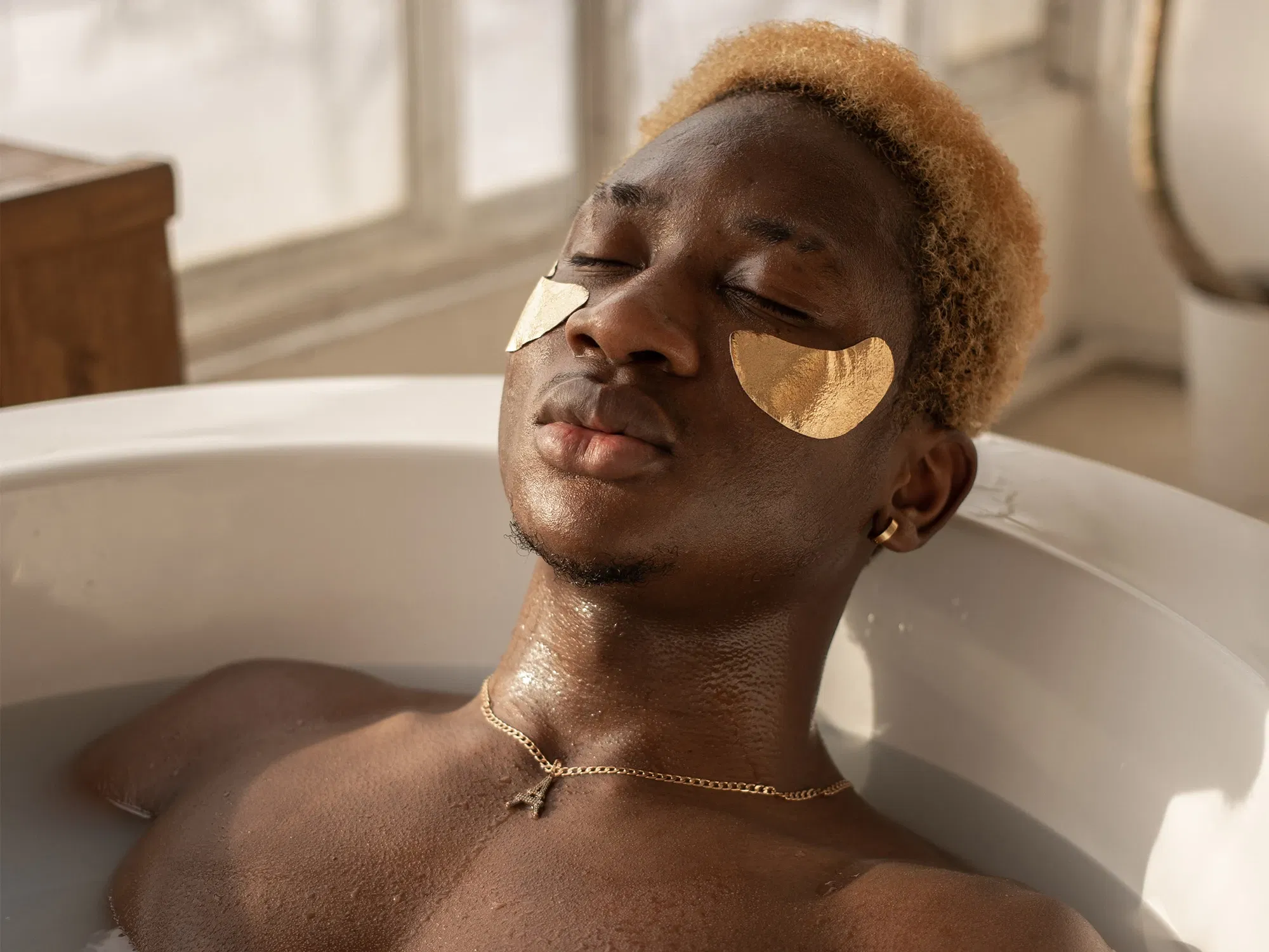 Ein Mann mit goldenen Augenpads liegt entspannt in einer mit Wasser gefüllten Badewanne. Sein Gesicht ist ruhig, seine Augen geschlossen, während weiches Tageslicht durch ein Fenster fällt und den Raum erhellt.