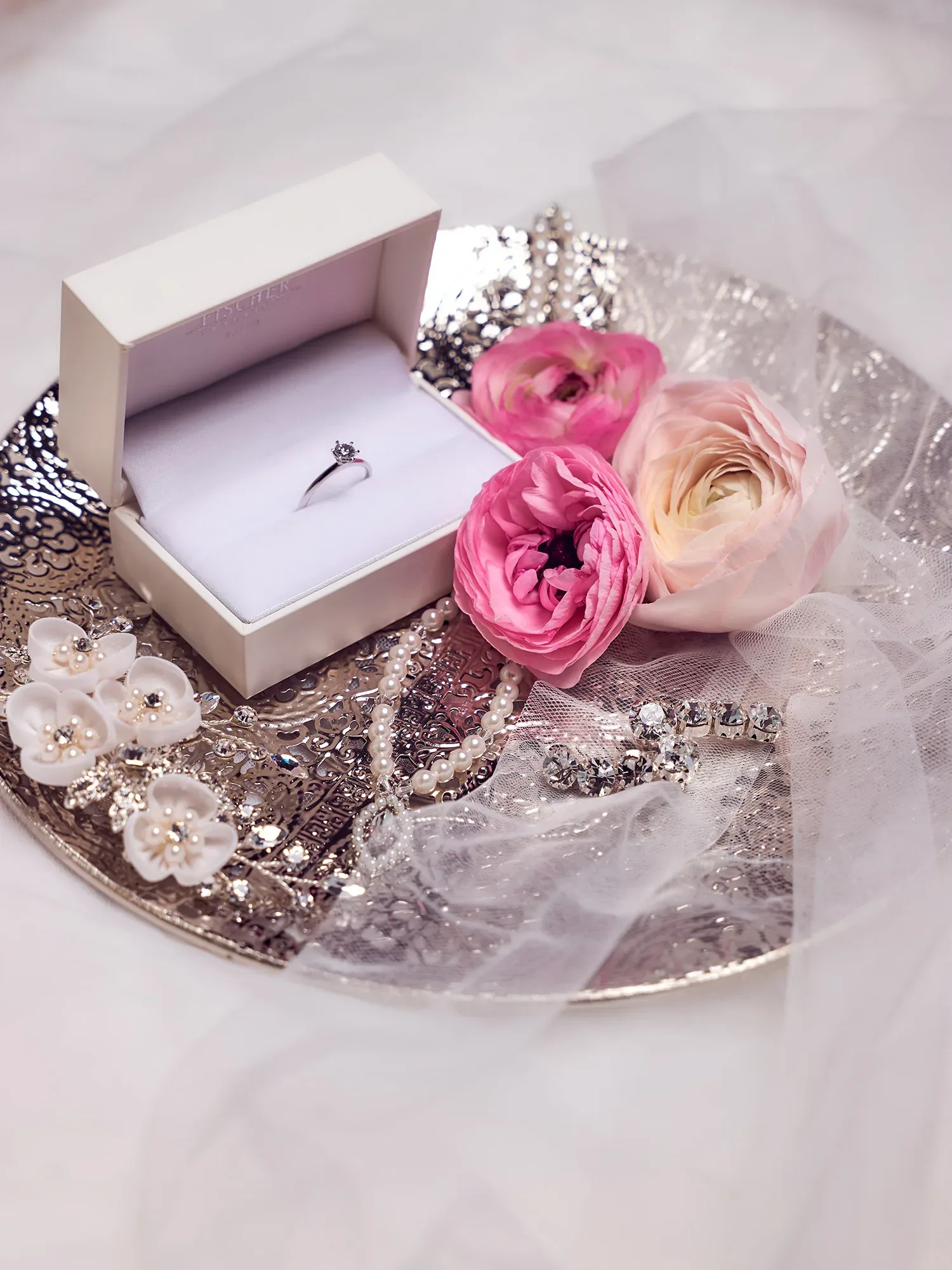 Auf einem silbernen Tablett liegt eine offene Schachtel mit einem Verlobungsring, daneben zarte rosa Ranunkeln, Perlen und glänzender Brautschmuck. Ein transparenter Schleier umrahmt das florale und elegante Arrangement, das eine feierliche Stimmung erzeugt.