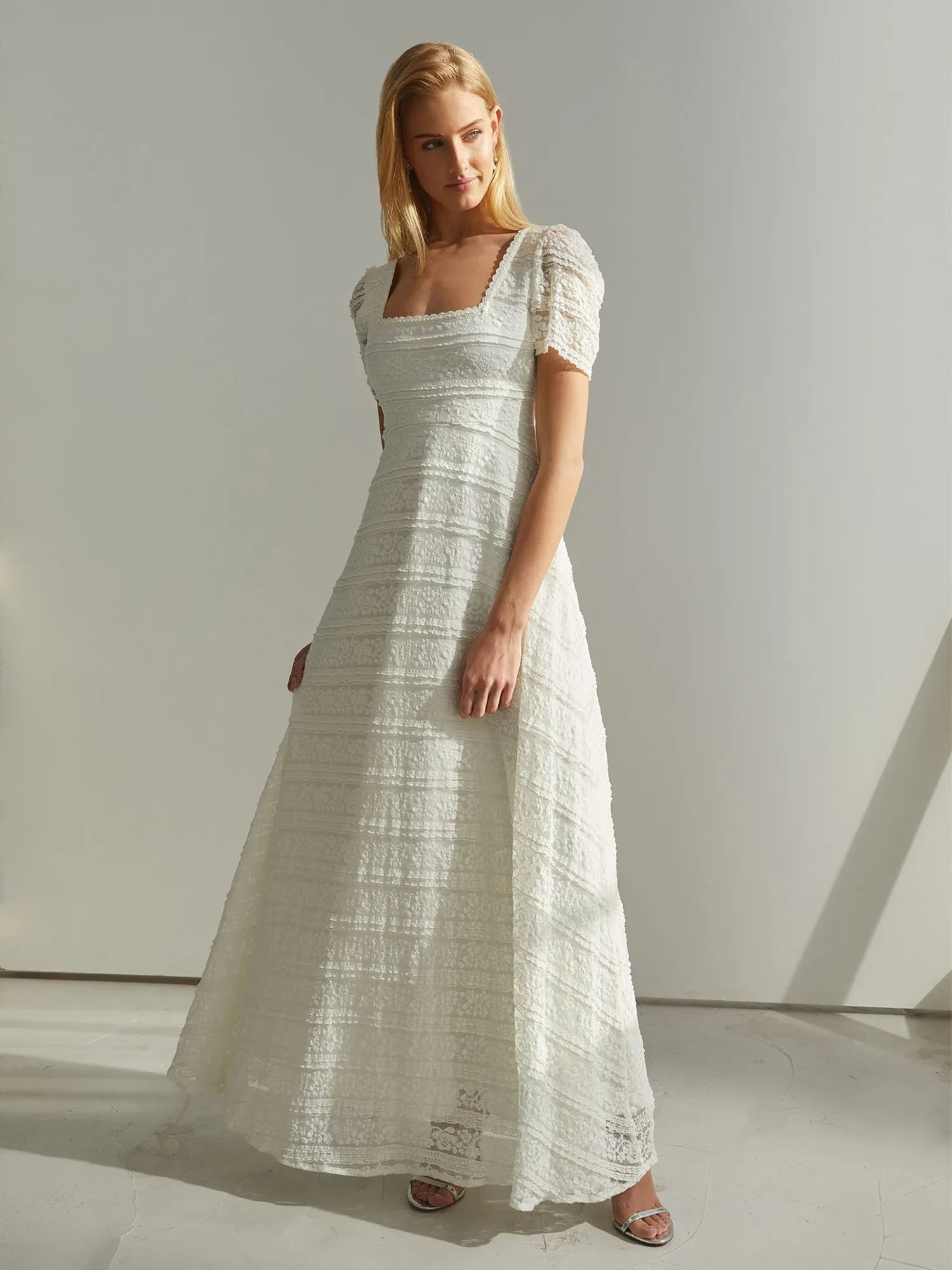 Eine Frau in einem eleganten, elfenbeinfarbenen Empire-Hochzeitskleid mit subtil strukturiertem Stoff und kurzen Puffärmeln steht in einem hellen, minimalistischen Raum. Das Kleid betont eine schlichte Eleganz mit seiner fließenden Silhouette und dem quadratischen Ausschnitt. Ihr Blick ist nachdenklich und abgewandt, während das sanfte Licht die feinen Details des Kleides hervorhebt und eine ruhige, sinnliche Atmosphäre schafft.