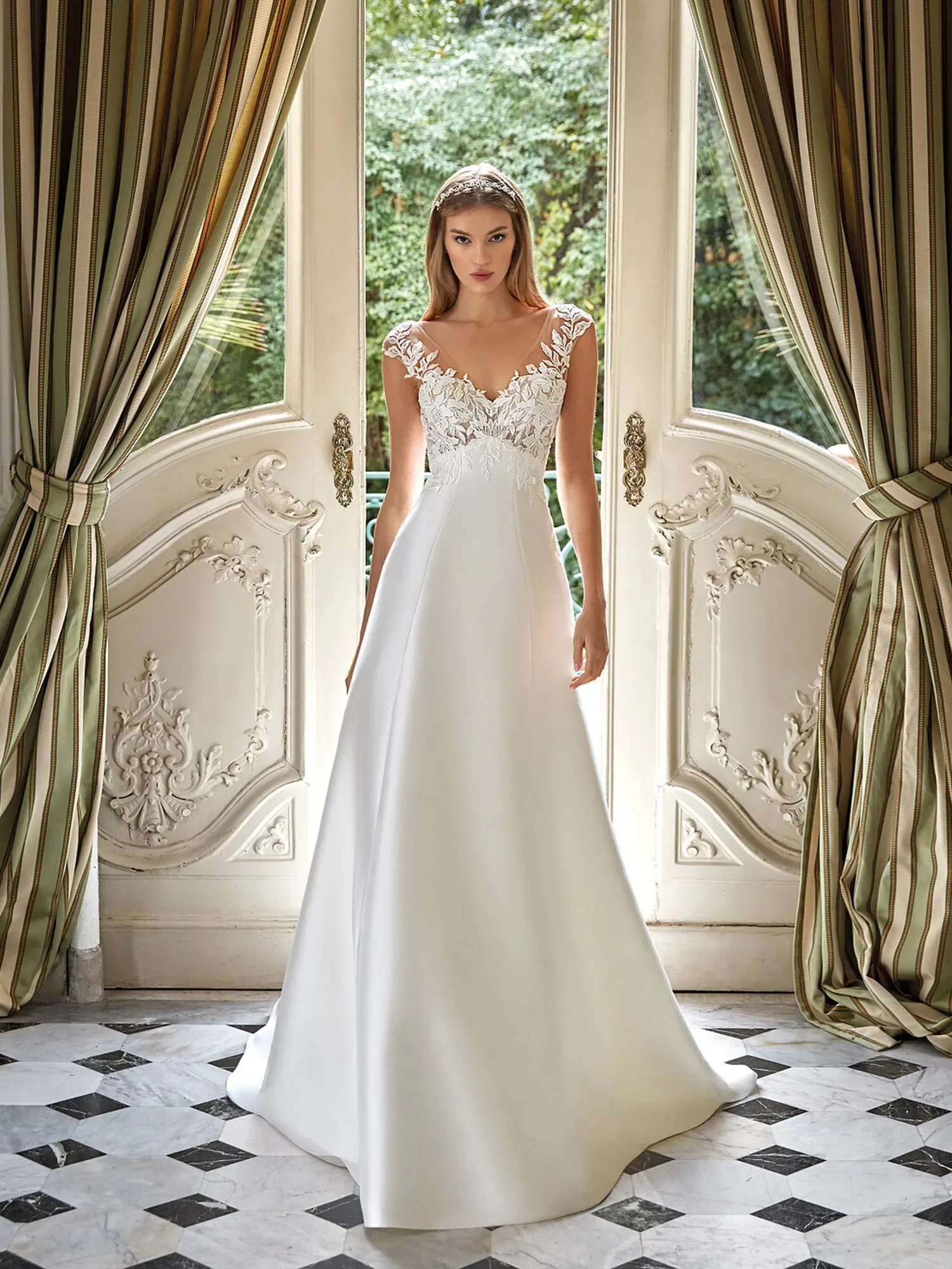 Eine Braut steht stolz in einem prachtvollen Empire-Hochzeitskleid mit zartem Spitzenoberteil und einer fließenden A-Linien-Rock aus Satin. Das Kleid hat einen V-Ausschnitt und ist in einem opulenten Raum mit hohen Fenstern und schweren gestreiften Vorhängen präsentiert. Der Boden ist mit Marmorfliesen in einem Schachbrettmuster geschmückt, was der Szene ein Gefühl von historischer Eleganz verleiht. Das sanfte Tageslicht betont die sorgfältigen Details des Spitzenoberteils und das strahlende Weiß des Rocks.