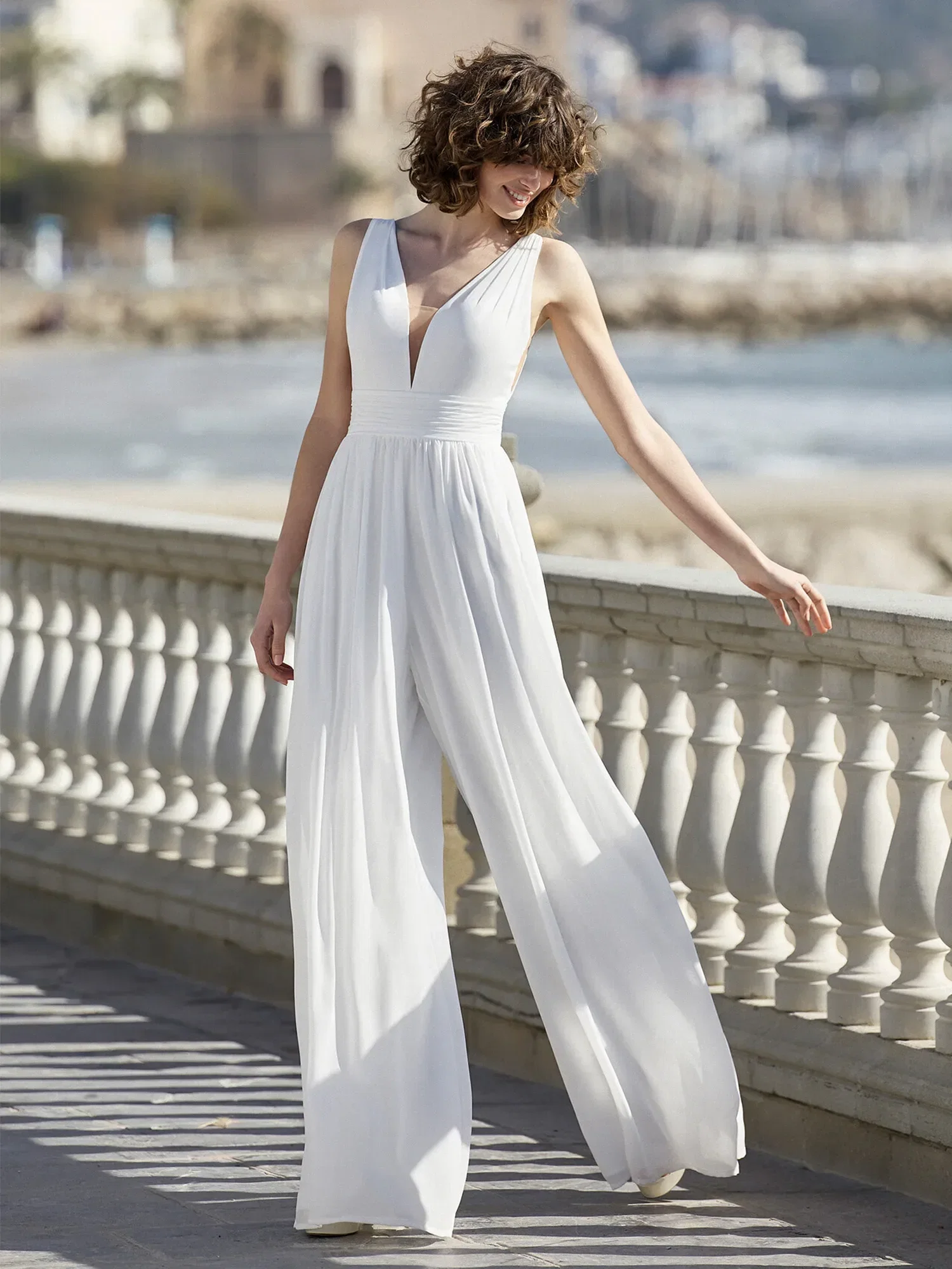 Eine Braut trägt einen weißen Jumpsuit mit weitem Bein und tiefem V-Ausschnitt. Sie posiert lächelnd auf einer Promenade am Meer und genießt den sanften Wind, der den fließenden Stoff ihres Outfits bewegt.