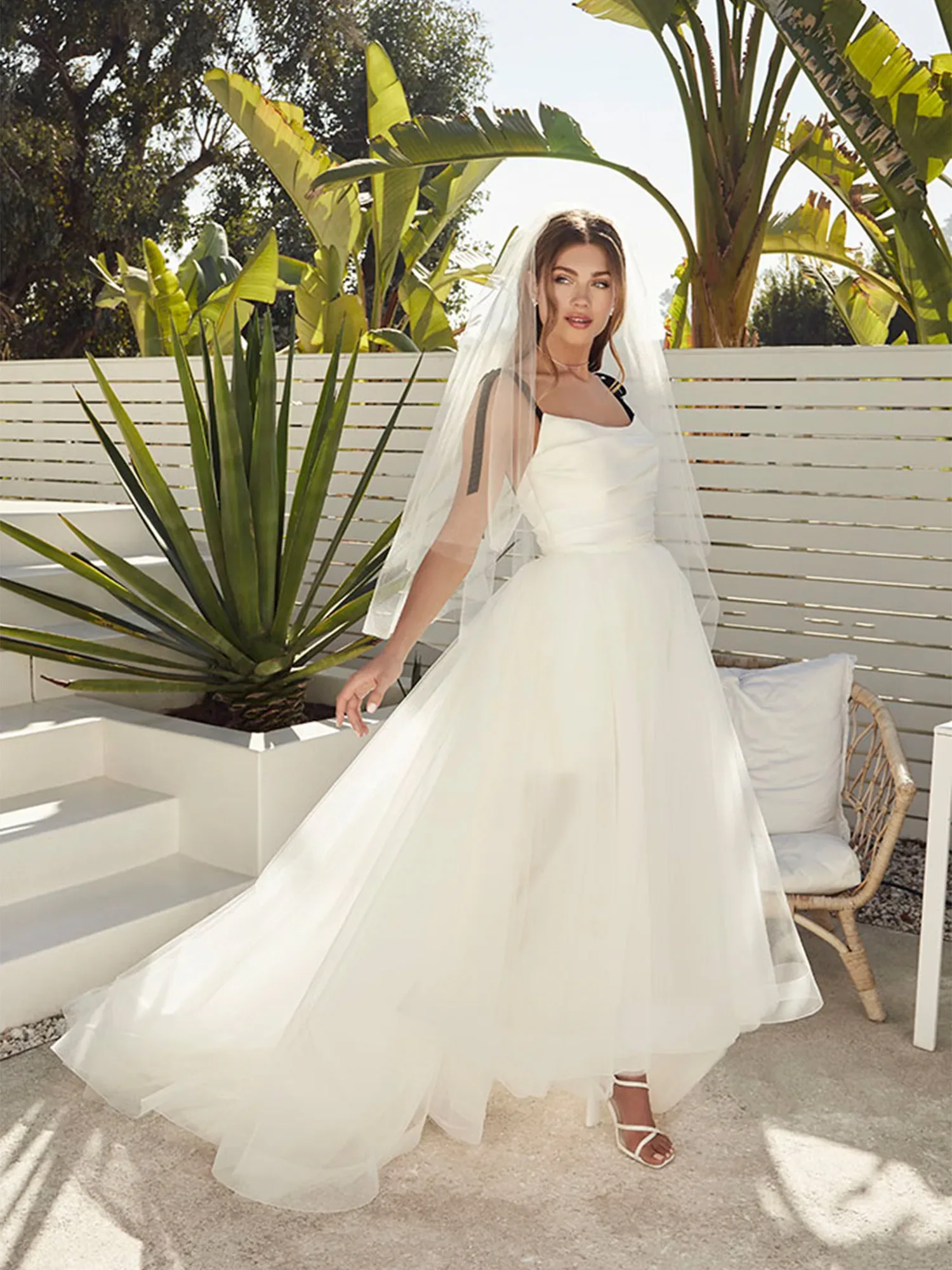 Eine Braut in einem modernen Hochzeitskleid mit Tüllrock posiert im Freien vor tropischen Pflanzen. Sie trägt einen langen Schleier, während der fließende Rock ihres Kleides elegant im Sonnenlicht weht.