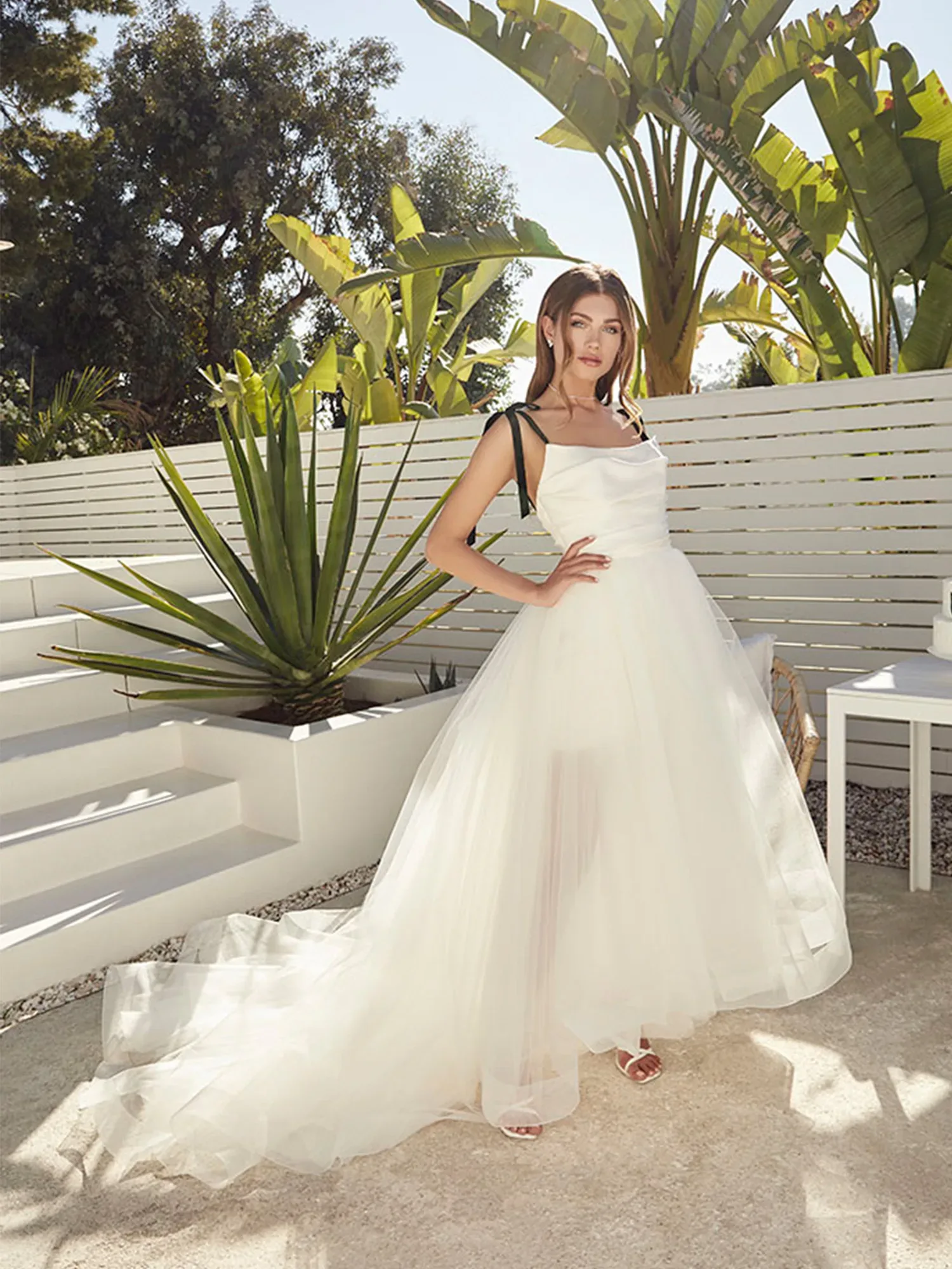 Eine Braut trägt ein modernes Hochzeitskleid mit asymmetrischem Tüllrock und posiert im Freien vor tropischen Pflanzen. Sie hat eine Hand an der Hüfte, während der fließende Rock ihres Kleides leicht vom Wind bewegt wird.