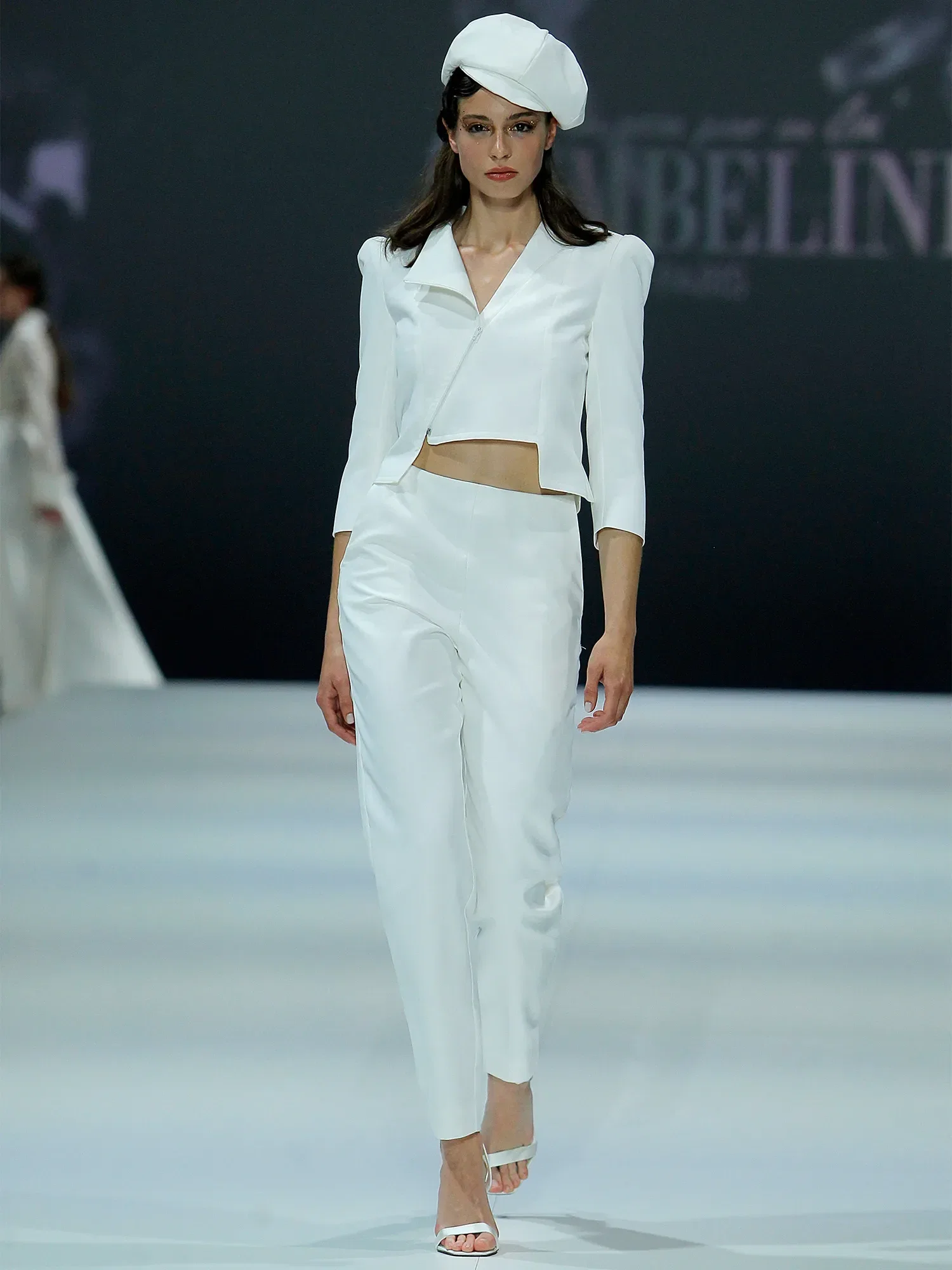 Eine Frau trägt ein modernes, zweiteiliges Brautoutfit mit einem kurzen Blazer und einer eleganten Hose. Sie geht über den Laufsteg und trägt dazu eine weiße, stilvolle Mütze. Das minimalistische Design des Outfits verleiht ihr einen eleganten und zeitgenössischen Look.