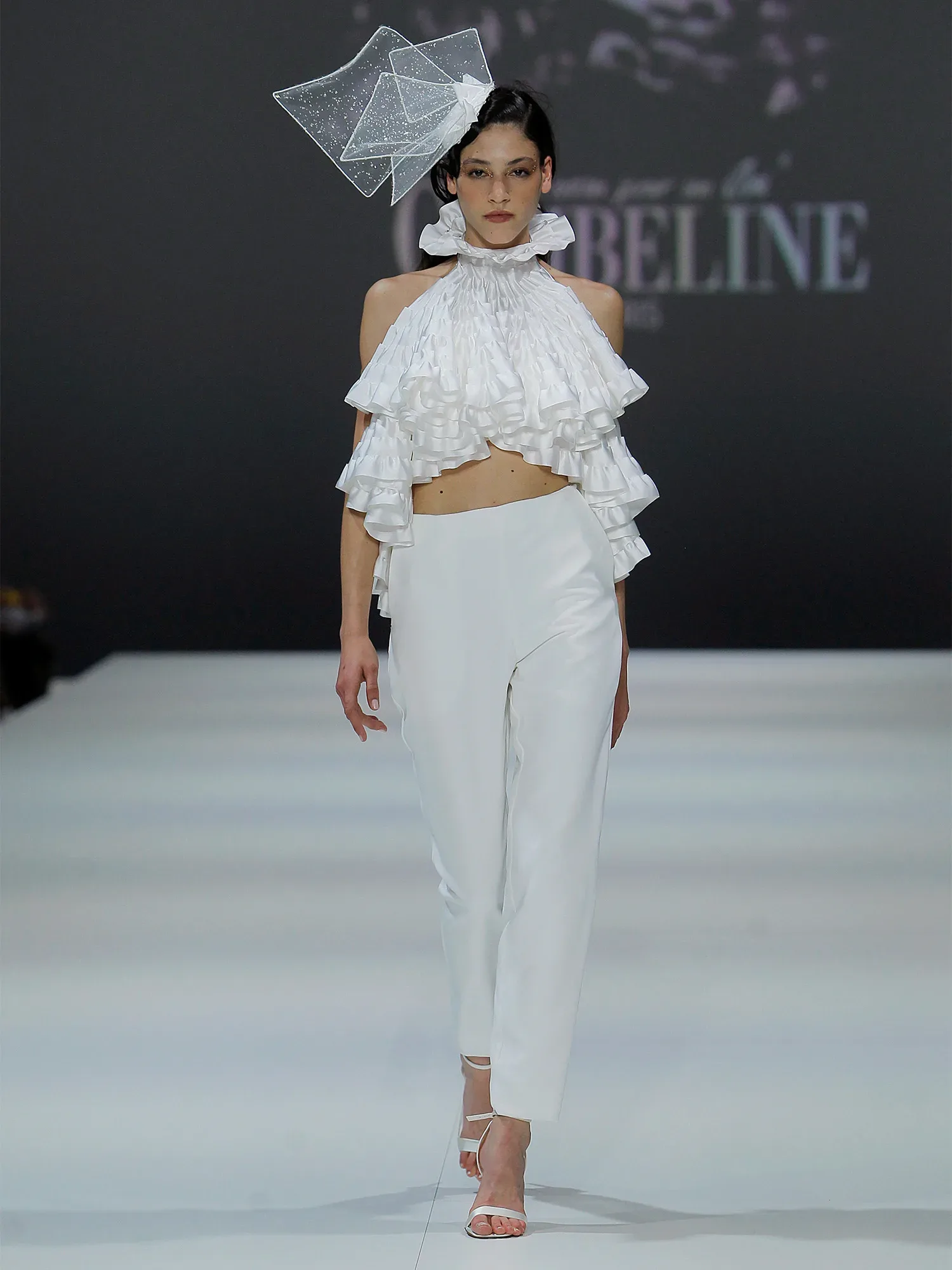 Ein Model präsentiert einen auffälligen Zweiteiler mit einem rüschenverzierten Oberteil und einer schmalen weißen Hose auf dem Laufsteg. Sie trägt dazu ein großes geometrisches Accessoire auf dem Kopf, das den avantgardistischen Look des Outfits betont.