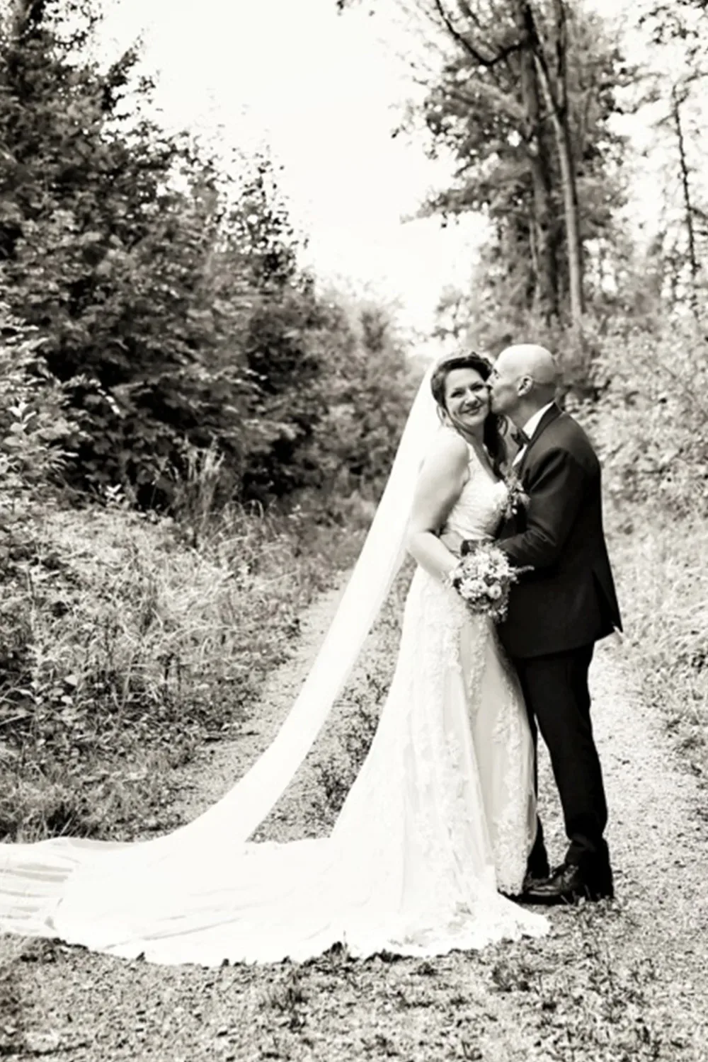 Schwarz-Weiß-Foto eines Hochzeitspaares, das auf einem Waldweg steht. Die Braut trägt ein Spitzenkleid mit langem Schleier und hält einen Blumenstrauß, während der Bräutigam im Anzug ihr einen Kuss auf die Wange gibt. Beide lächeln glücklich in die Kamera, umgeben von Natur.