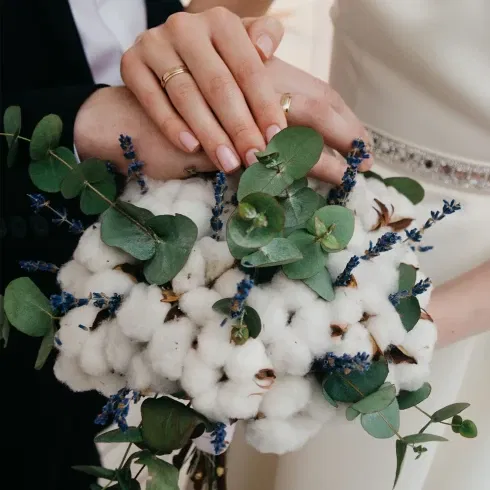 Ein romantisches Bild eines Brautpaares mit zarten Eheringen, die über einem Strauß aus Baumwolle, Eukalyptus und Lavendel ruhen.