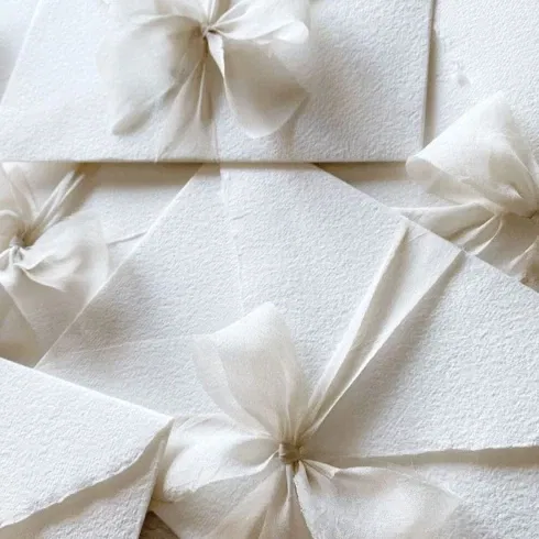Eine Nahaufnahme von mehreren weißen Umschlägen, die jeweils mit zarten, durchsichtigen Schleifen aus Organza verziert sind. Die Umschläge liegen übereinander und erzeugen ein elegantes, festliches Muster.