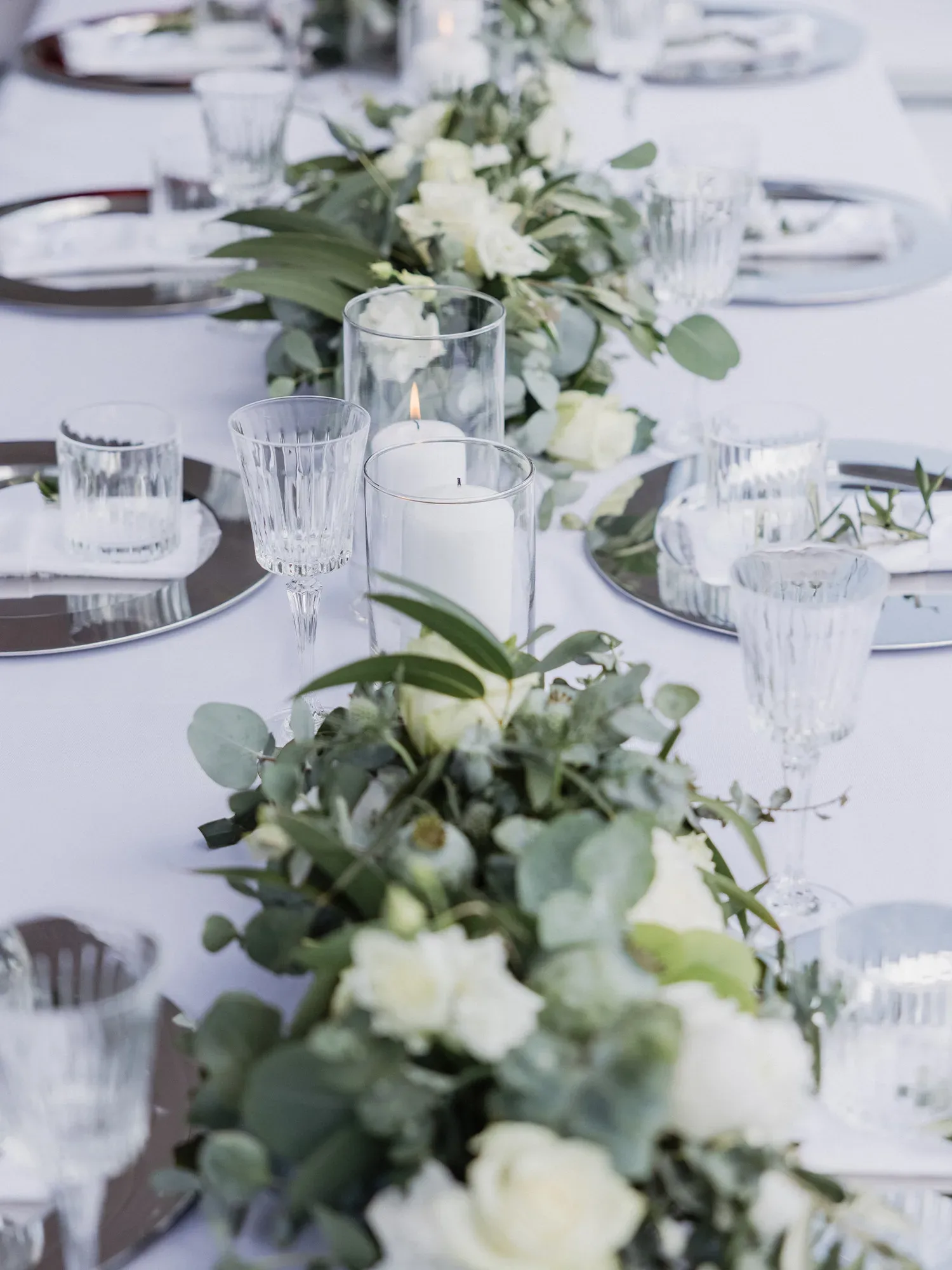 Eine festliche Hochzeitstafel mit eleganter Tischdekoration. Eukalyptusblätter und weiße Rosen schmücken die Mitte des Tisches, umgeben von stilvollen Kerzen in Glasgefäßen. Ein Brautstrauß aus frischem Eukalyptus und weißen Blumen, gehalten von der Braut im Hintergrund eines weiten Feldes.