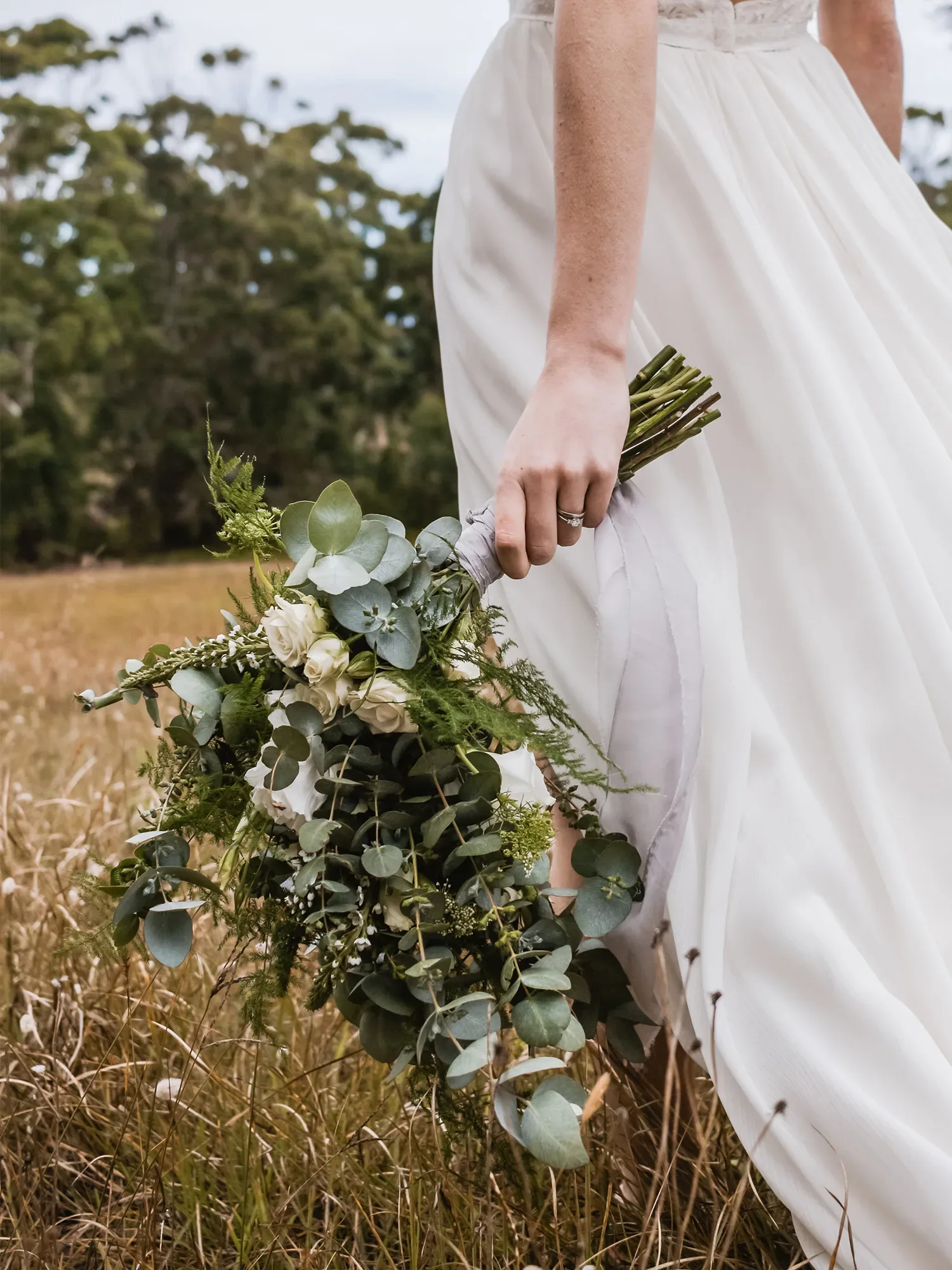Eine Braut hält einen üppigen Brautstrauß aus Eukalyptus, Olivenzweigen und weißen Blumen, während sie durch ein Feld in ihrem Hochzeitskleid schreitet.