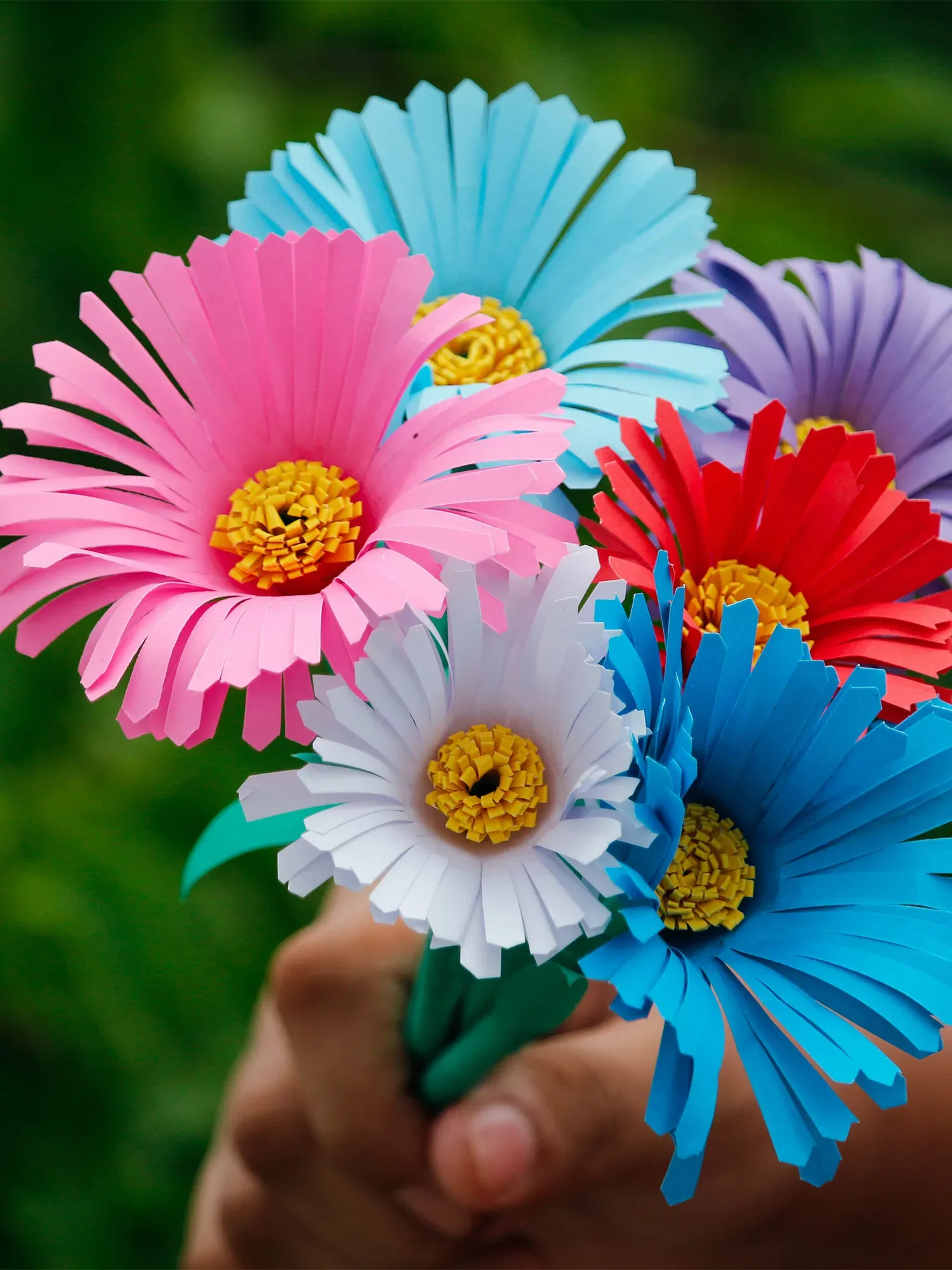 Ein bunter Strauß aus handgefertigten Papierblumen in lebhaften Farben wie Pink, Blau, Rot und Weiß, gehalten von einer Hand im Vordergrund.