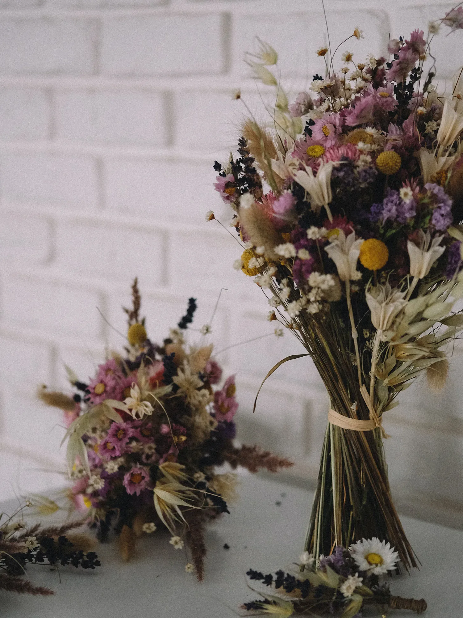Ein großer, bunter Trockenblumenstrauß aus Lavendel, Gräsern und Blumen, arrangiert vor einer weißen Backsteinwand.