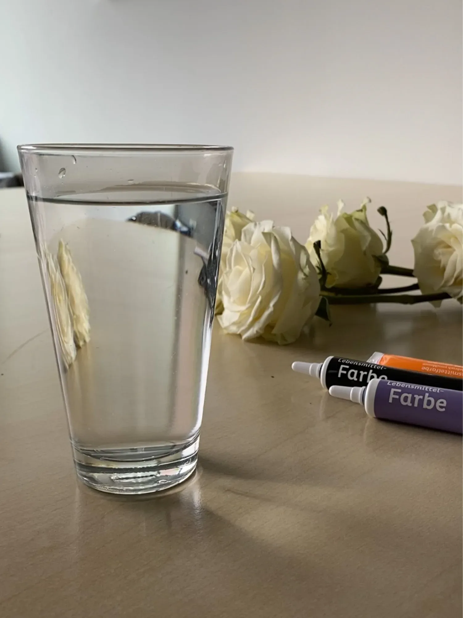 Ein Glas Wasser steht auf einem Tisch, daneben liegen mehrere weiße Rosen und Tuben mit Lebensmittelfarbe in verschiedenen Farben. Das Glas und die Blumen spiegeln sich leicht auf der Tischoberfläche wider.