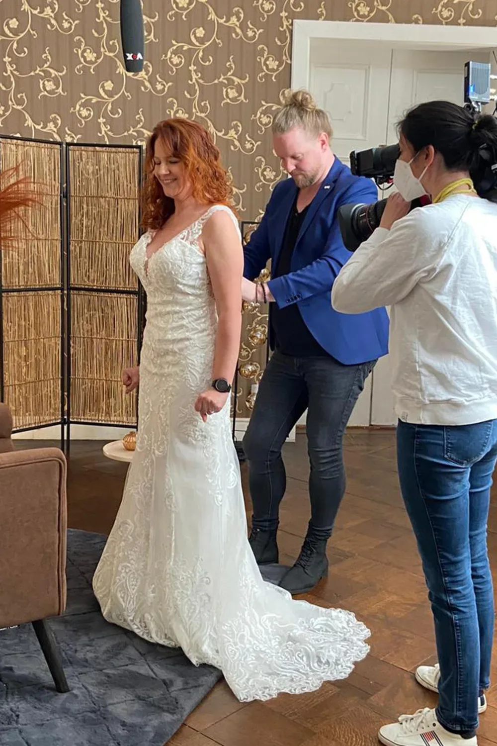 Eine Frau in einem eleganten weißen Brautkleid wird von einem Mann im blauen Blazer am Rücken des Kleides angepasst. Eine Kamerafrau filmt die Szene, während ein Mikrofon von oben hängt. Der Raum ist stilvoll mit einer gemusterten Tapete und modernen Dekorationen gestaltet.