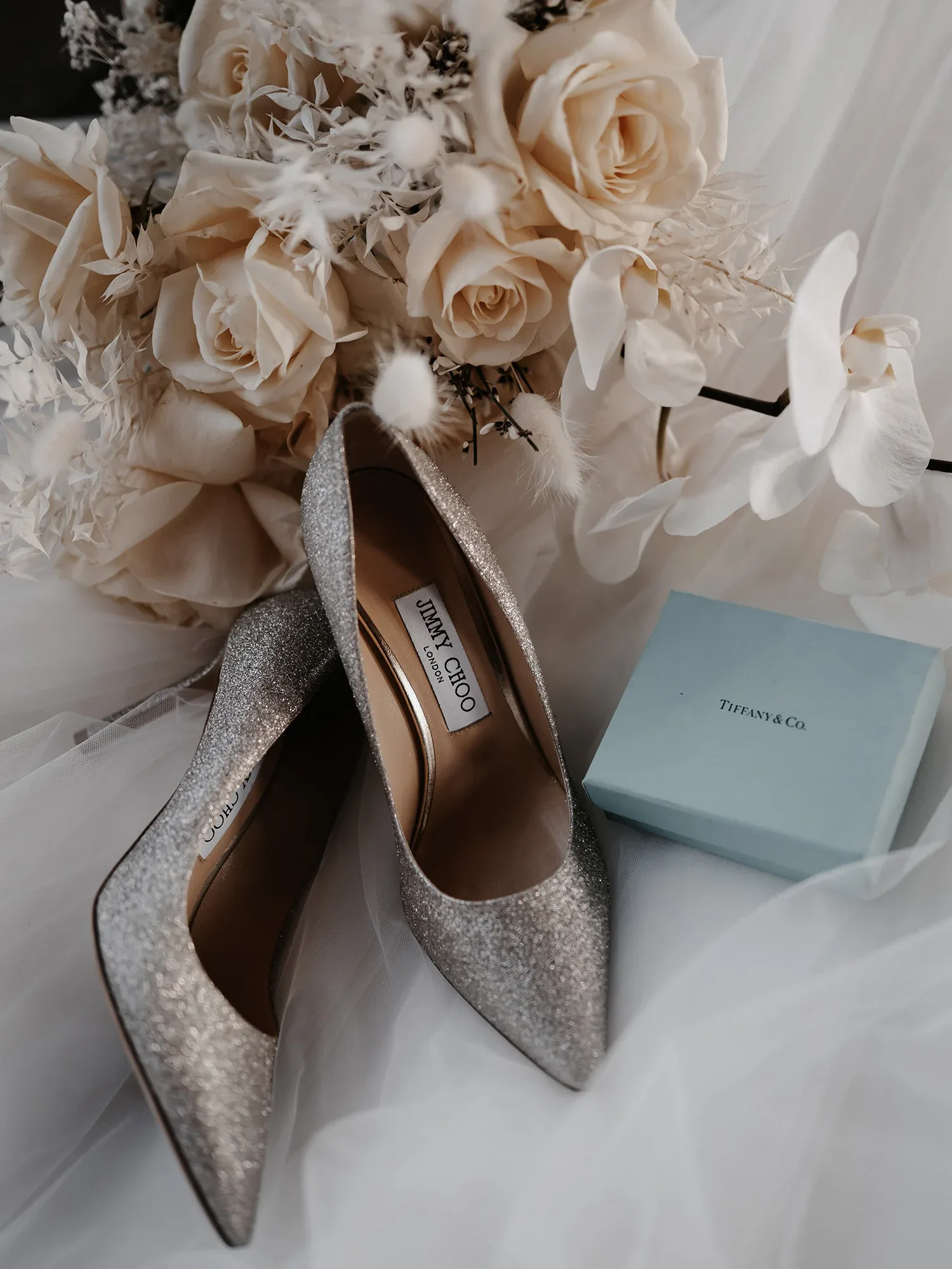 Elegante, silberne Jimmy Choo Pumps liegen neben einem Brautstrauß aus cremefarbenen Rosen und einer Tiffany & Co. Box auf weißem Tüll.