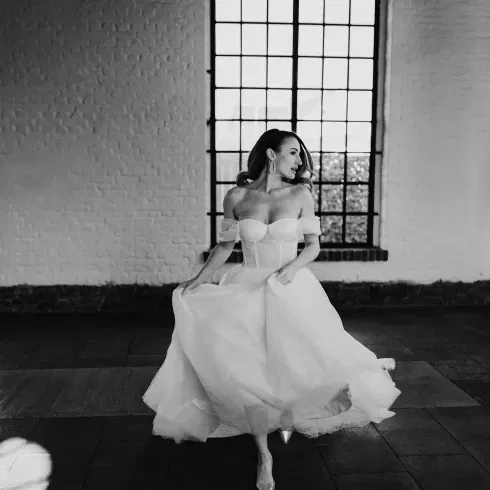 Eine Braut in einem modernen, trägerlosen Kleid tanzt in einem Raum mit hohen, gewölbten Fenstern und wirft einen eleganten Blick über die Schulter.