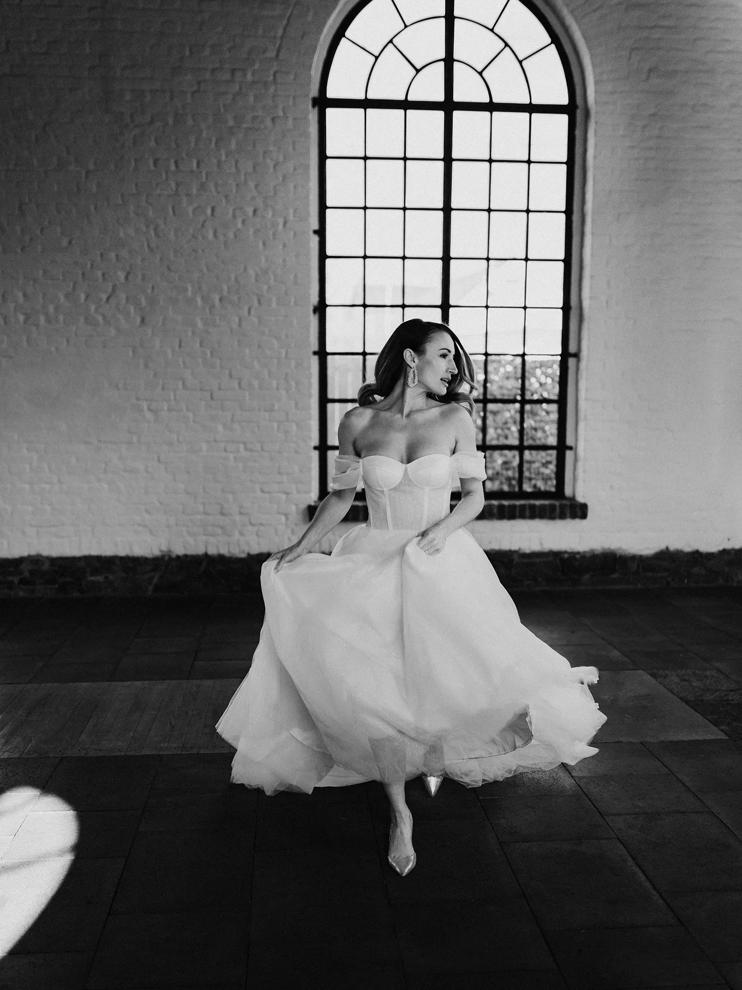 Eine Braut in einem modernen, trägerlosen Kleid tanzt in einem Raum mit hohen, gewölbten Fenstern und wirft einen eleganten Blick über die Schulter.