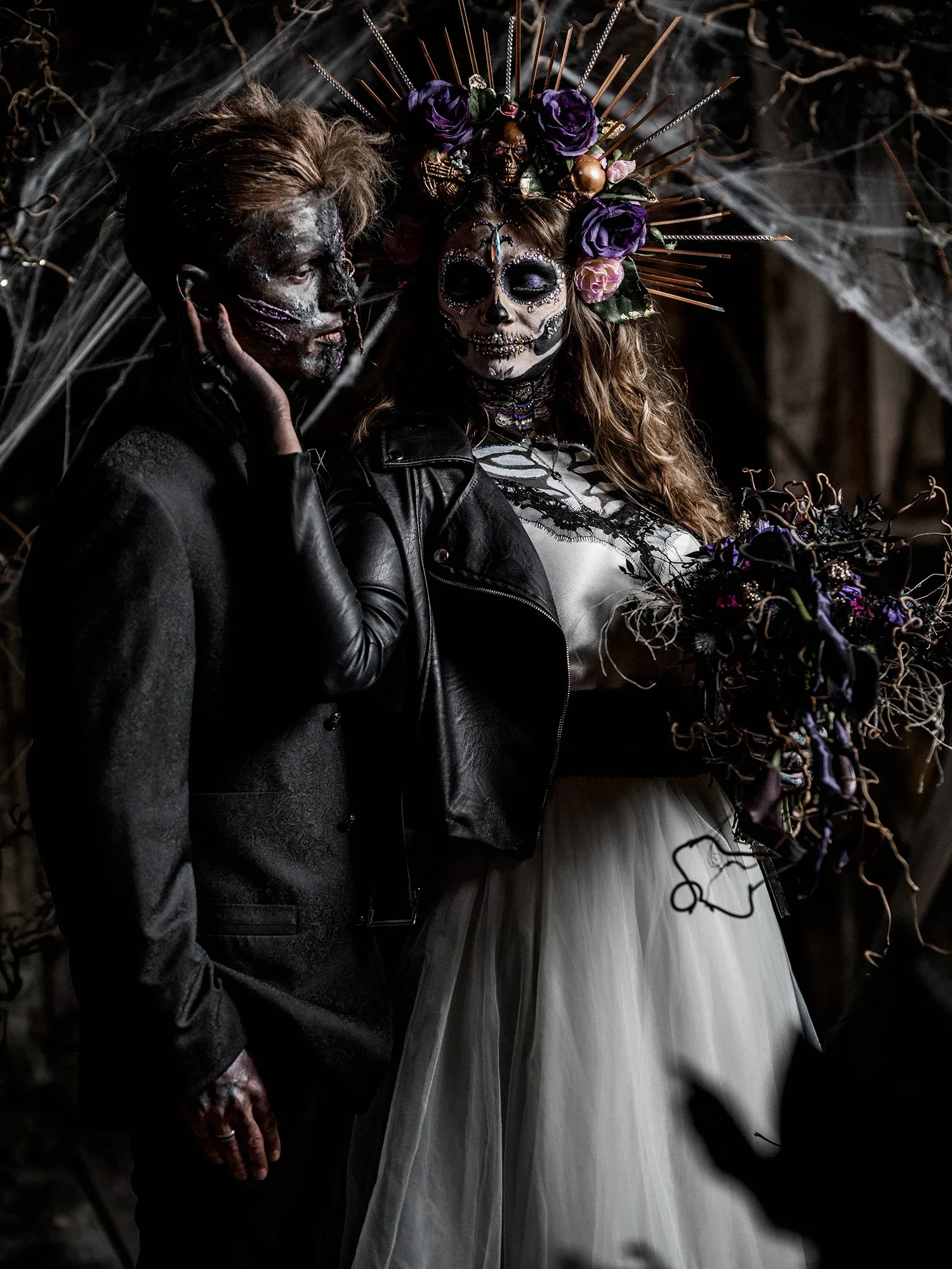 Ein Brautpaar in Gothic-Hochzeitskleidung, das sich an einem düsteren, spinnennetzverzierten Ort befindet. Beide tragen dunkles, dramatisches Make-up und Blumenkränze.