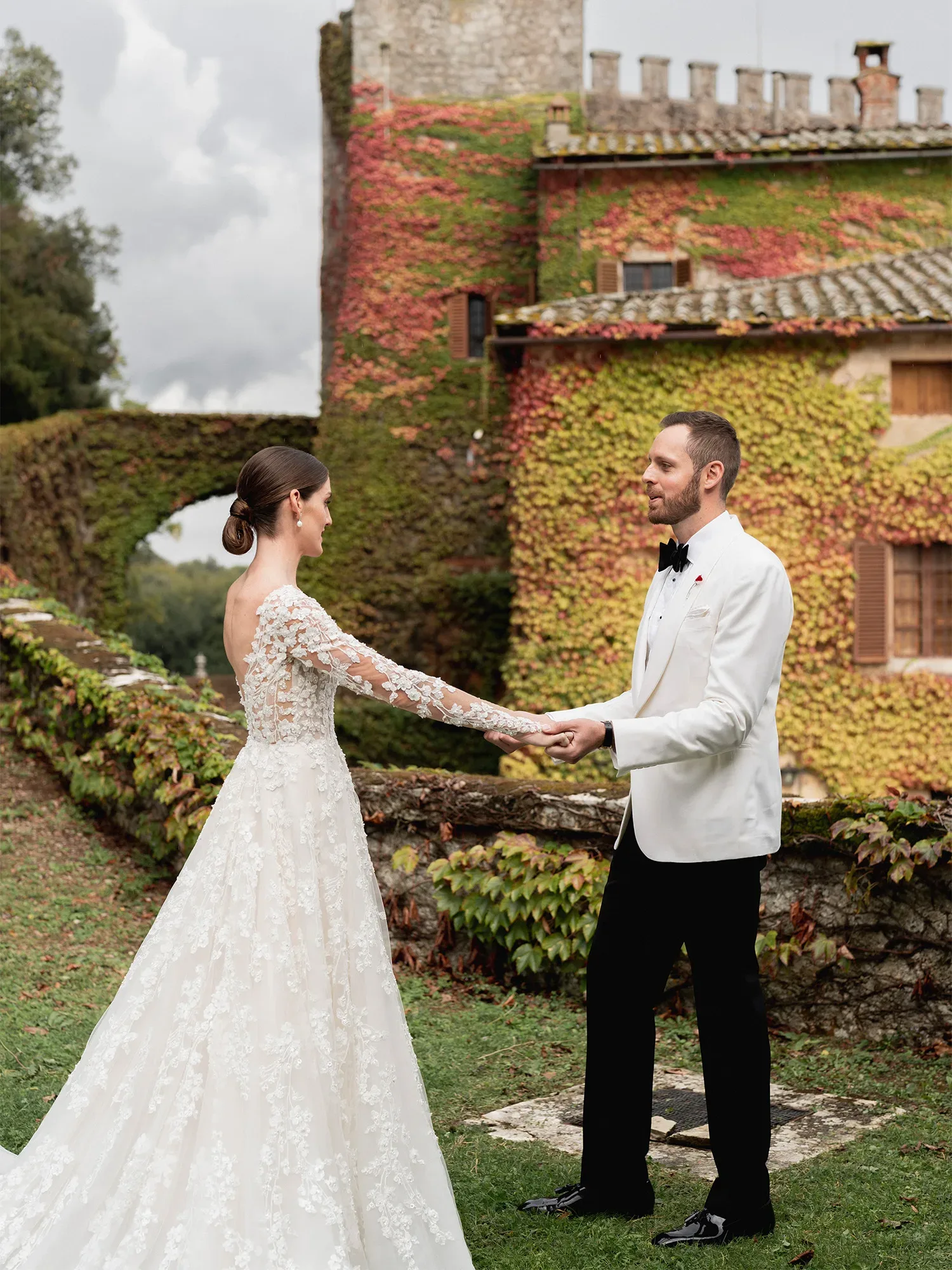 Ein Hochzeitspaar steht sich auf dem Rasen eines historischen Anwesens gegenüber und hält sich an den Händen. Die Braut trägt ein langes Spitzenkleid mit transparenten Ärmeln, während der Bräutigam einen weißen Smoking trägt.