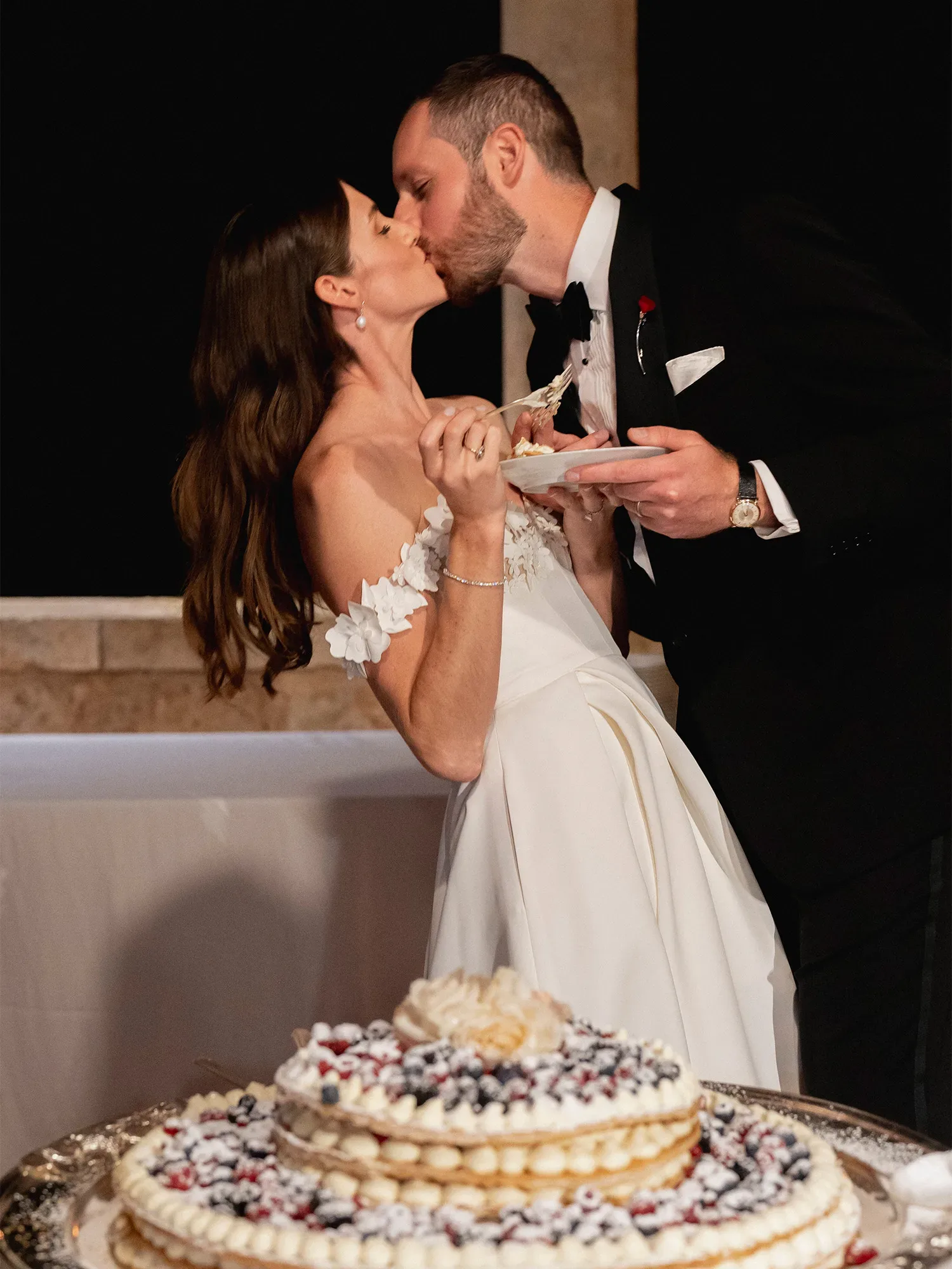 Ein Hochzeitspaar teilt einen Kuss während des Anschnitts ihrer Hochzeitstorte. Die Braut hält ein Stück Kuchen auf einem Teller, während der Bräutigam ebenfalls einen Teller in der Hand hält. Im Vordergrund ist eine mehrstöckige Torte mit Beeren und Sahne zu sehen.