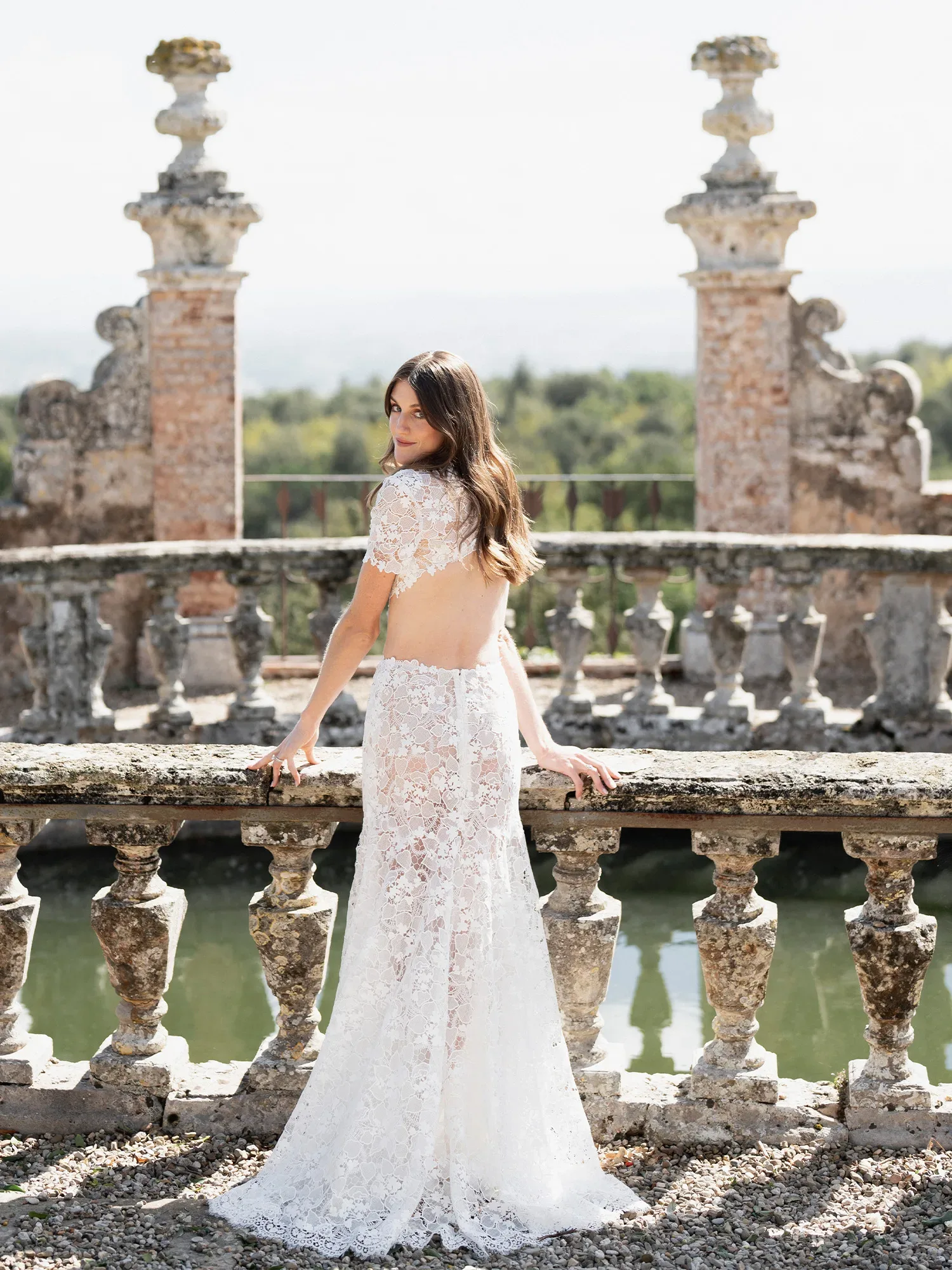 Eine Braut in einem rückenfreien, durchsichtigen Spitzenkleid lehnt sich an eine alte steinerne Balustrade mit Blick auf einen Teich. Sie blickt über ihre Schulter zurück und lächelt sanft in die Kamera.