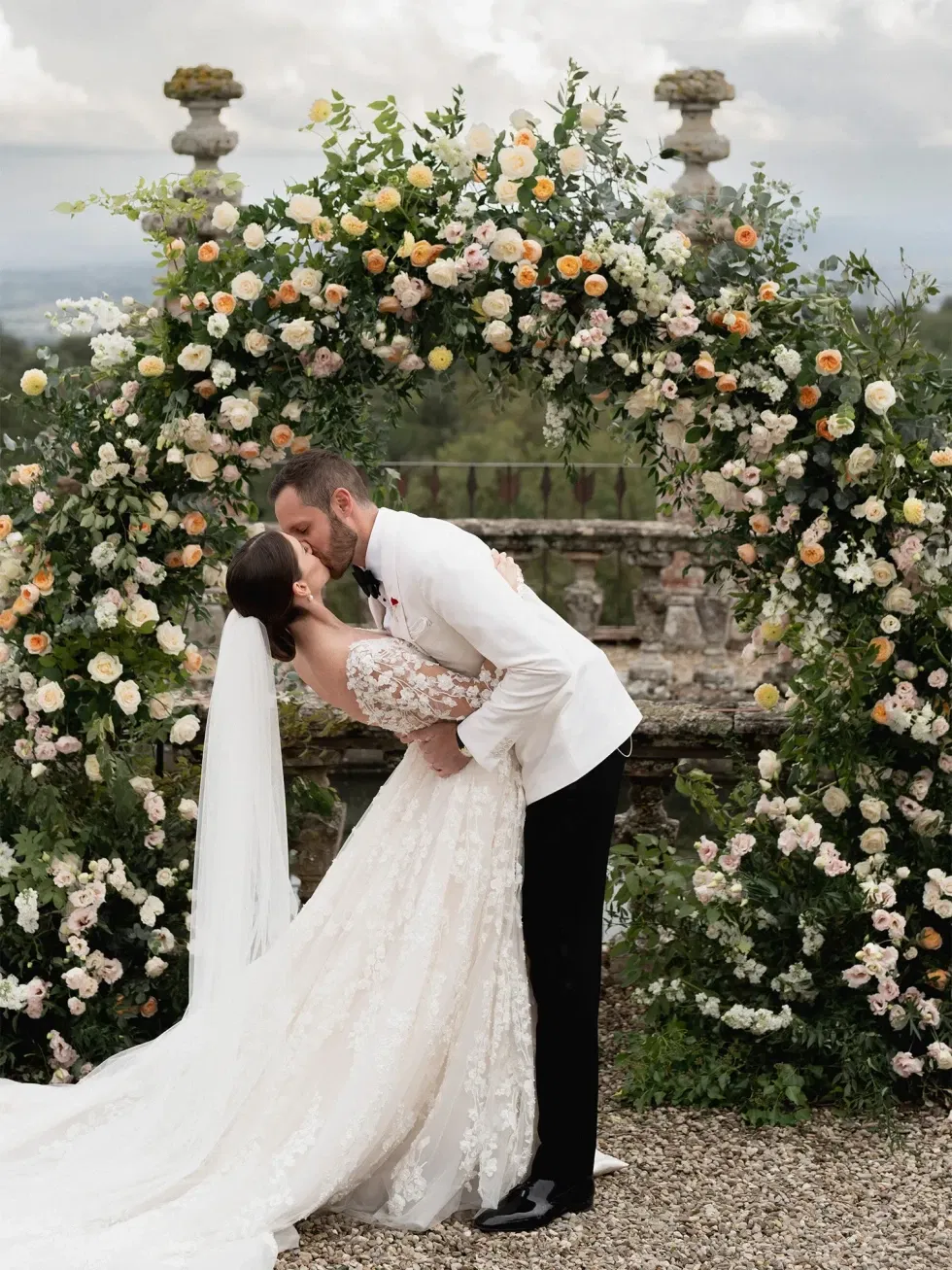 Ein Hochzeitspaar teilt einen romantischen Kuss unter einem üppigen Blumenbogen, umgeben von Rosen und Grünpflanzen. Die Braut trägt ein aufwendig verziertes Spitzenkleid, während der Bräutigam einen weißen Smoking trägt.