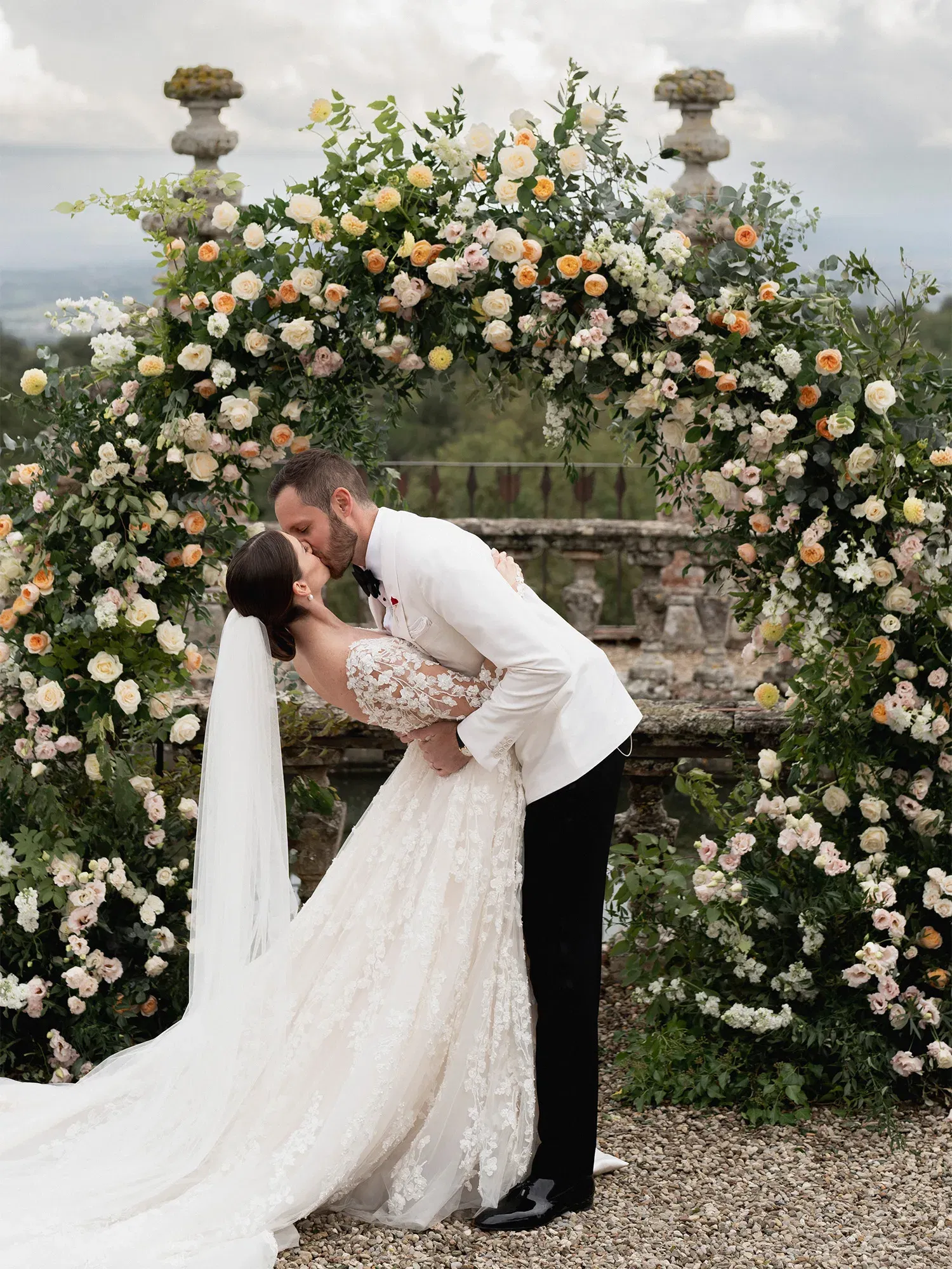 Ein Hochzeitspaar teilt einen romantischen Kuss unter einem üppigen Blumenbogen, umgeben von Rosen und Grünpflanzen. Die Braut trägt ein aufwendig verziertes Spitzenkleid, während der Bräutigam einen weißen Smoking trägt.