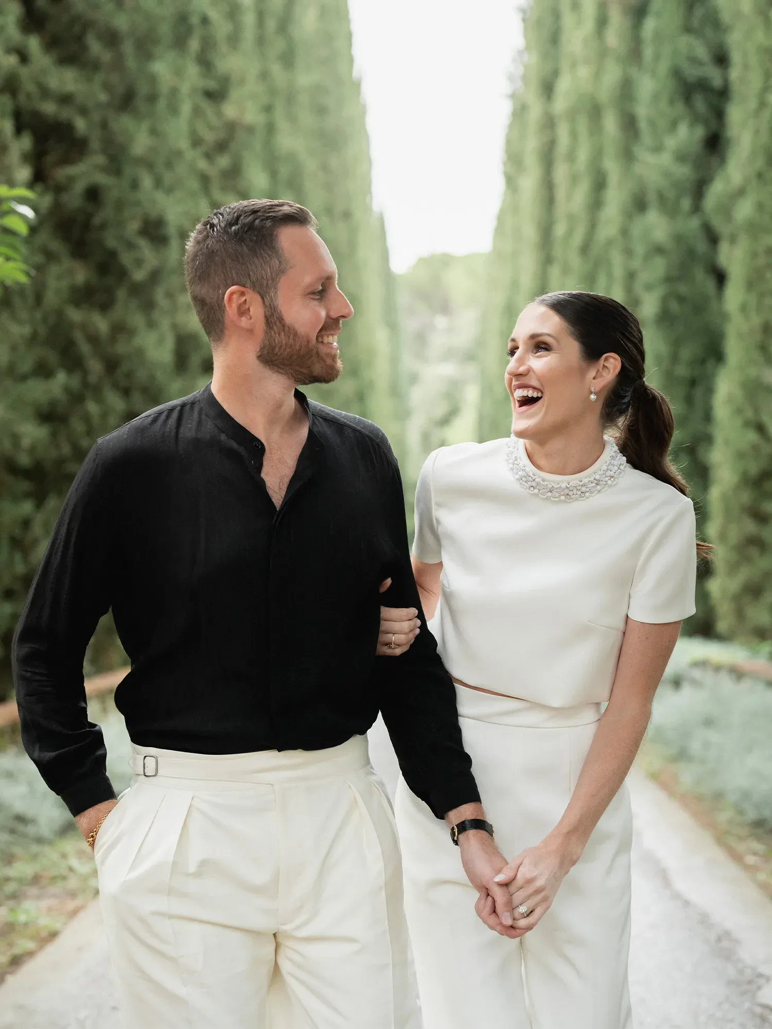 Ein lächelndes Paar spaziert Arm in Arm einen von hohen Bäumen gesäumten Weg entlang. Der Mann trägt ein schwarzes Hemd und weiße Hosen, die Frau ein schlichtes, elegantes weißes Ensemble und lacht fröhlich.
