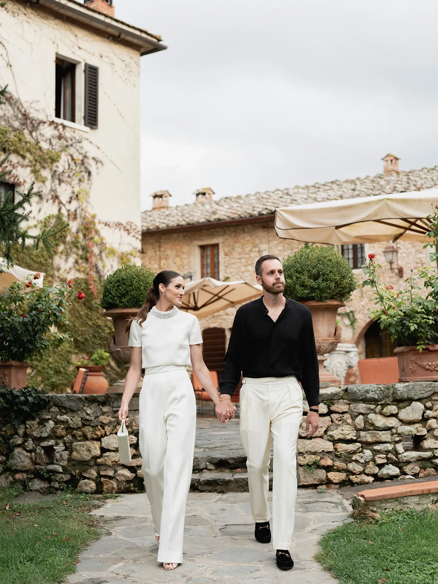 Ein stilvolles Paar spaziert Hand in Hand durch den Garten eines rustikalen Anwesens. Die Frau trägt ein weißes, elegantes Ensemble und eine kleine Handtasche, während der Mann ein schwarzes Hemd und weiße Hosen trägt.