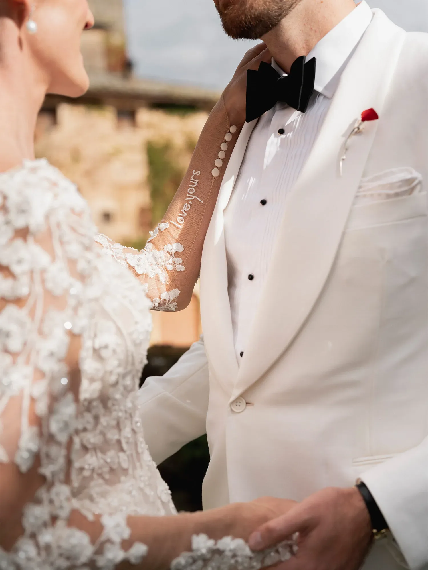 Nahaufnahme eines Hochzeitspaares, das sich liebevoll an den Händen hält. Der Bräutigam trägt einen weißen Smoking mit schwarzer Fliege, die Braut ein kunstvoll verziertes Spitzenkleid. Auf dem transparenten Ärmel der Braut ist der Schriftzug 'Love, yours' zu sehen.