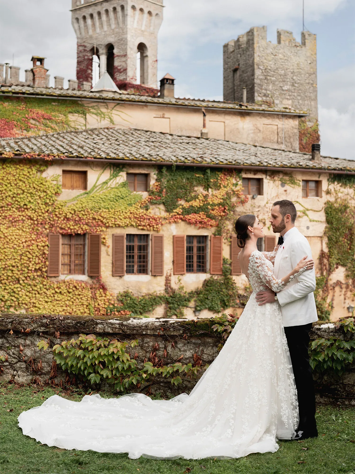 Ein Hochzeitspaar steht sich liebevoll gegenüber vor einer mit Efeu bedeckten, historischen Villa. Die Braut trägt ein florales Spitzenkleid mit langer Schleppe, während der Bräutigam einen weißen Smoking mit schwarzer Fliege trägt.