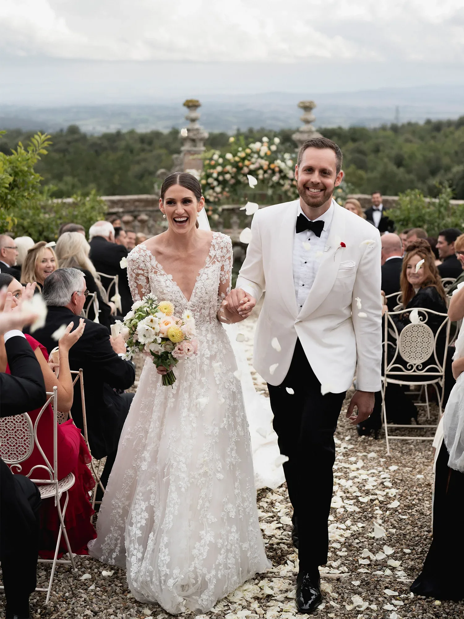 Ein strahlendes Hochzeitspaar schreitet nach der Zeremonie den Gang entlang, umgeben von applaudierenden Gästen. Die Braut trägt ein langes Spitzenkleid mit Blumenstrauß in der Hand, während der Bräutigam einen weißen Smoking trägt.
