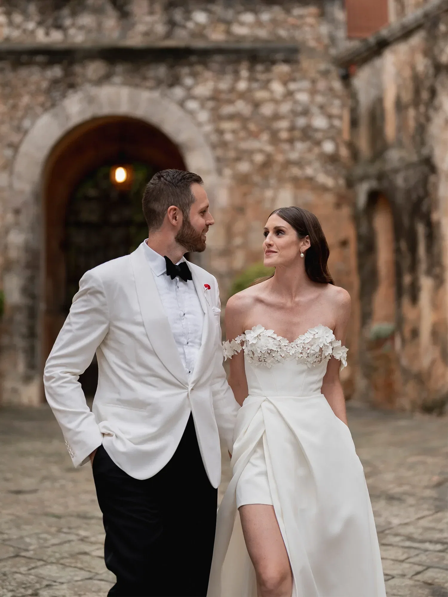 Ein Hochzeitspaar spaziert Hand in Hand durch einen historischen Innenhof. Die Braut trägt ein schulterfreies Kleid mit floralen Details und hohem Beinschlitz, während der Bräutigam einen weißen Smoking mit schwarzer Fliege trägt.