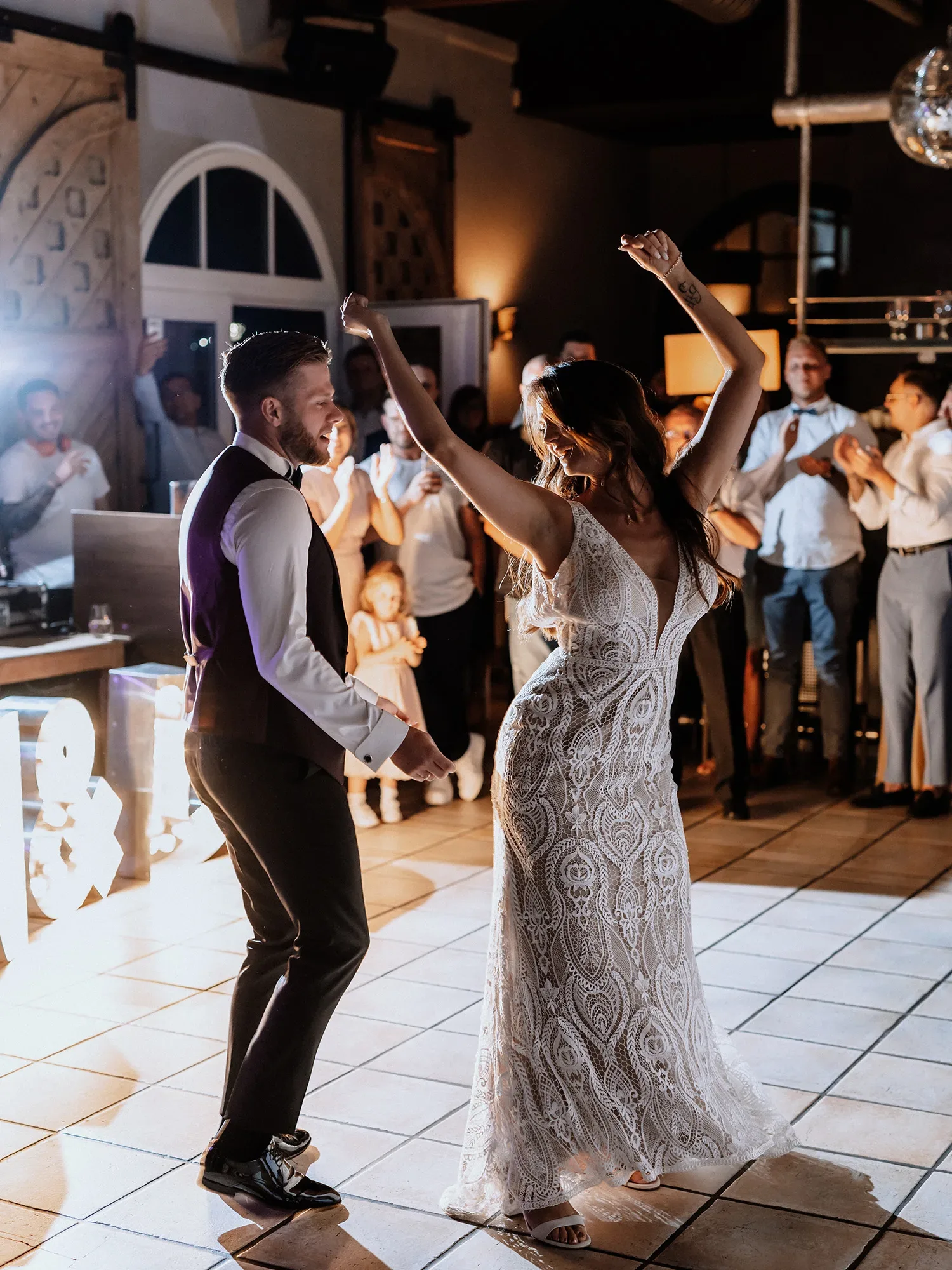 Braut und Bräutigam tanzen fröhlich auf der Tanzfläche während ihrer Hochzeit, umgeben von applaudierenden Gästen. Die Braut trägt ein detailliertes Spitzenkleid, während der Bräutigam einen eleganten Anzug mit Weste trägt.