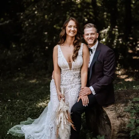 Ein glückliches Hochzeitspaar sitzt in einem Wald auf einem Baumstumpf. Die Braut trägt ein weißes, detailliertes Spitzenkleid, während der Bräutigam einen dunklen Anzug trägt. Beide lächeln und wirken entspannt
