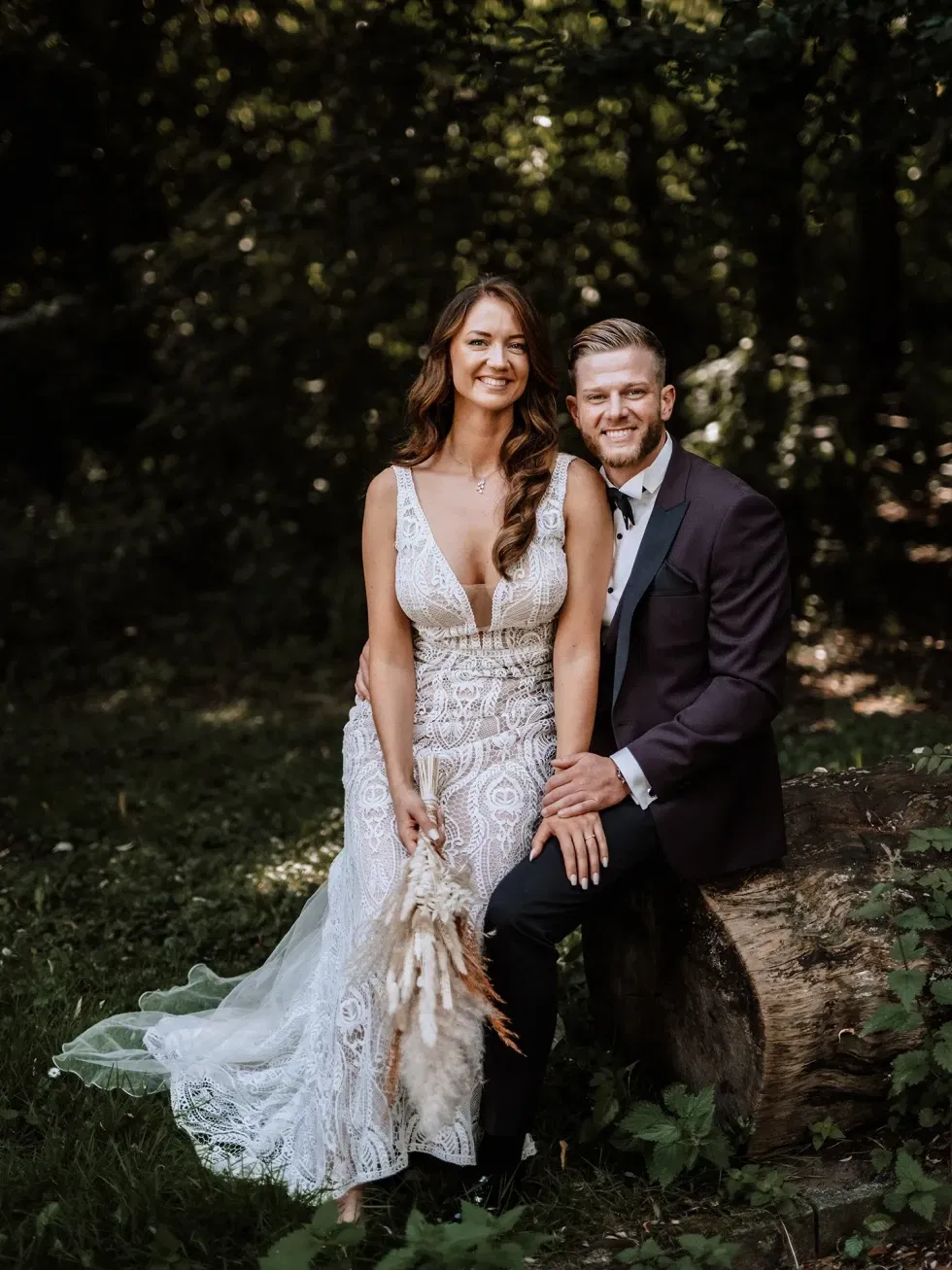 Ein glückliches Hochzeitspaar sitzt in einem Wald auf einem Baumstumpf. Die Braut trägt ein weißes, detailliertes Spitzenkleid, während der Bräutigam einen dunklen Anzug trägt. Beide lächeln und wirken entspannt