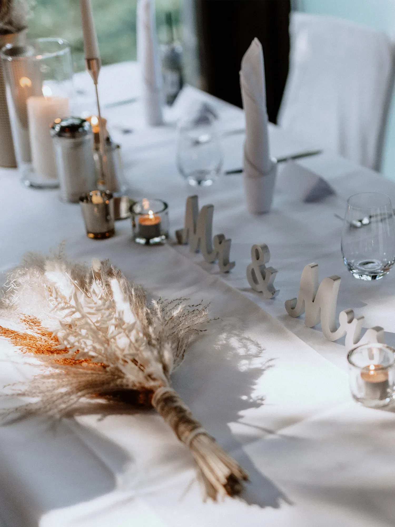 Detailansicht einer Hochzeitstafel mit einem Strauß aus getrockneten Gräsern und der Aufschrift 'Mr & Mrs' als Dekoration. Die Tischdekoration ist elegant und minimalistisch, mit Kerzen und gefalteten Servietten.