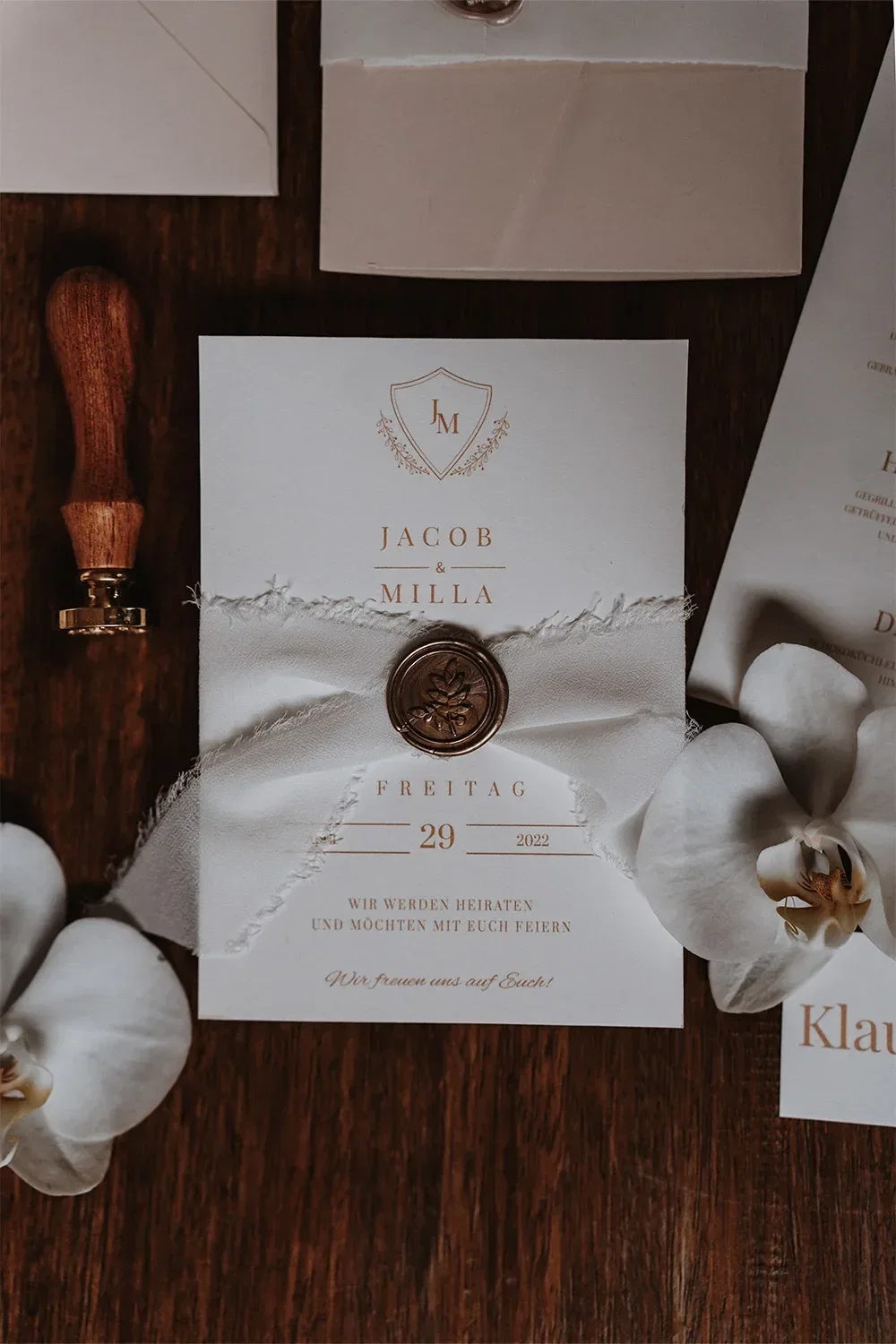 Eine elegante Hochzeitseinladung mit goldener Schrift und einem Wachssiegel, verziert mit weißem Band und Orchideenblüten auf einem Holztisch.
