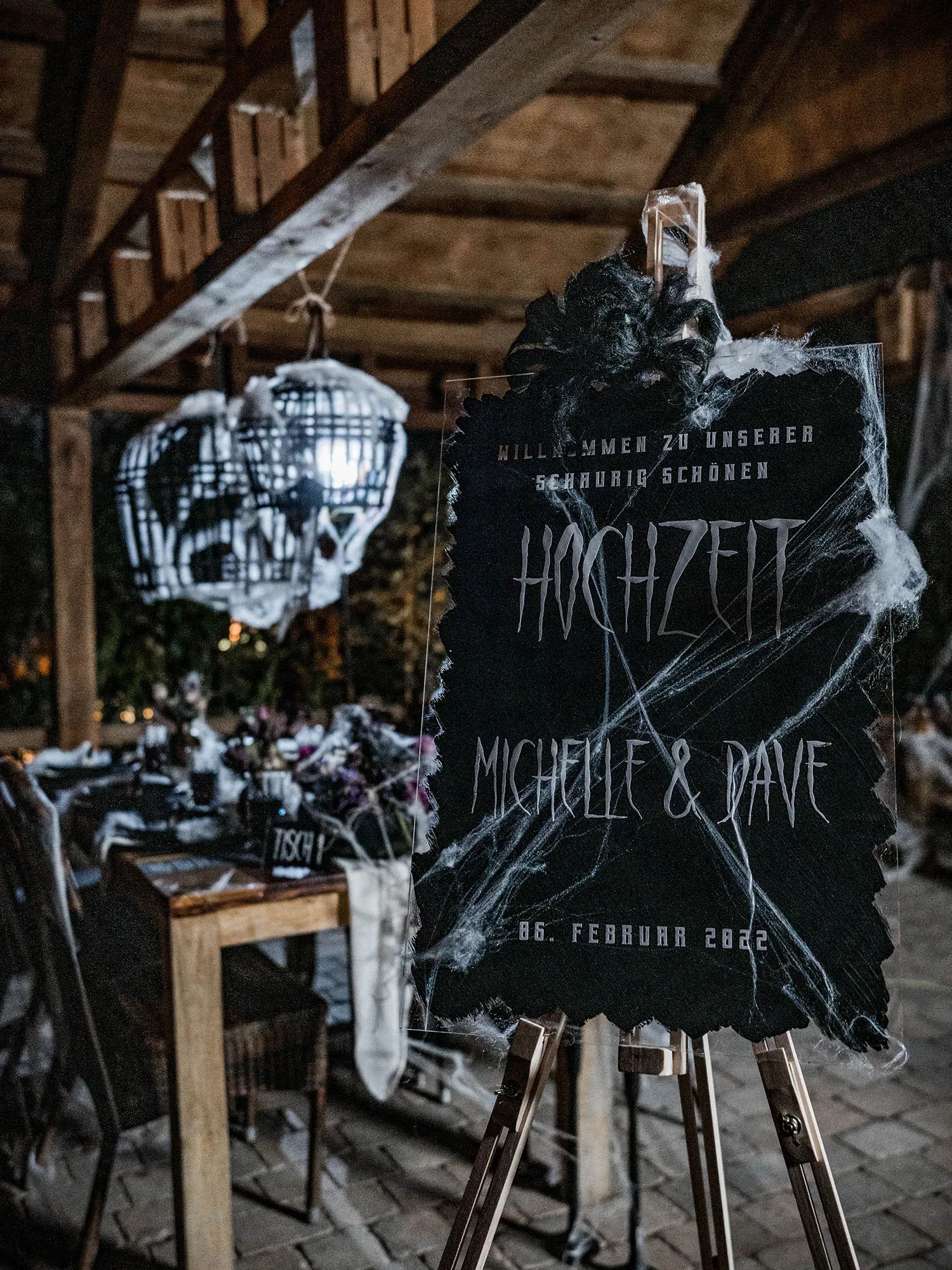 Ein schwarzes Hochzeitsschild mit Spinnweben, das die Namen "Michelle & Dave" und das Hochzeitsdatum zeigt. Im Hintergrund steht ein gedeckter Tisch in einem düsteren, stimmungsvollen Ambiente.