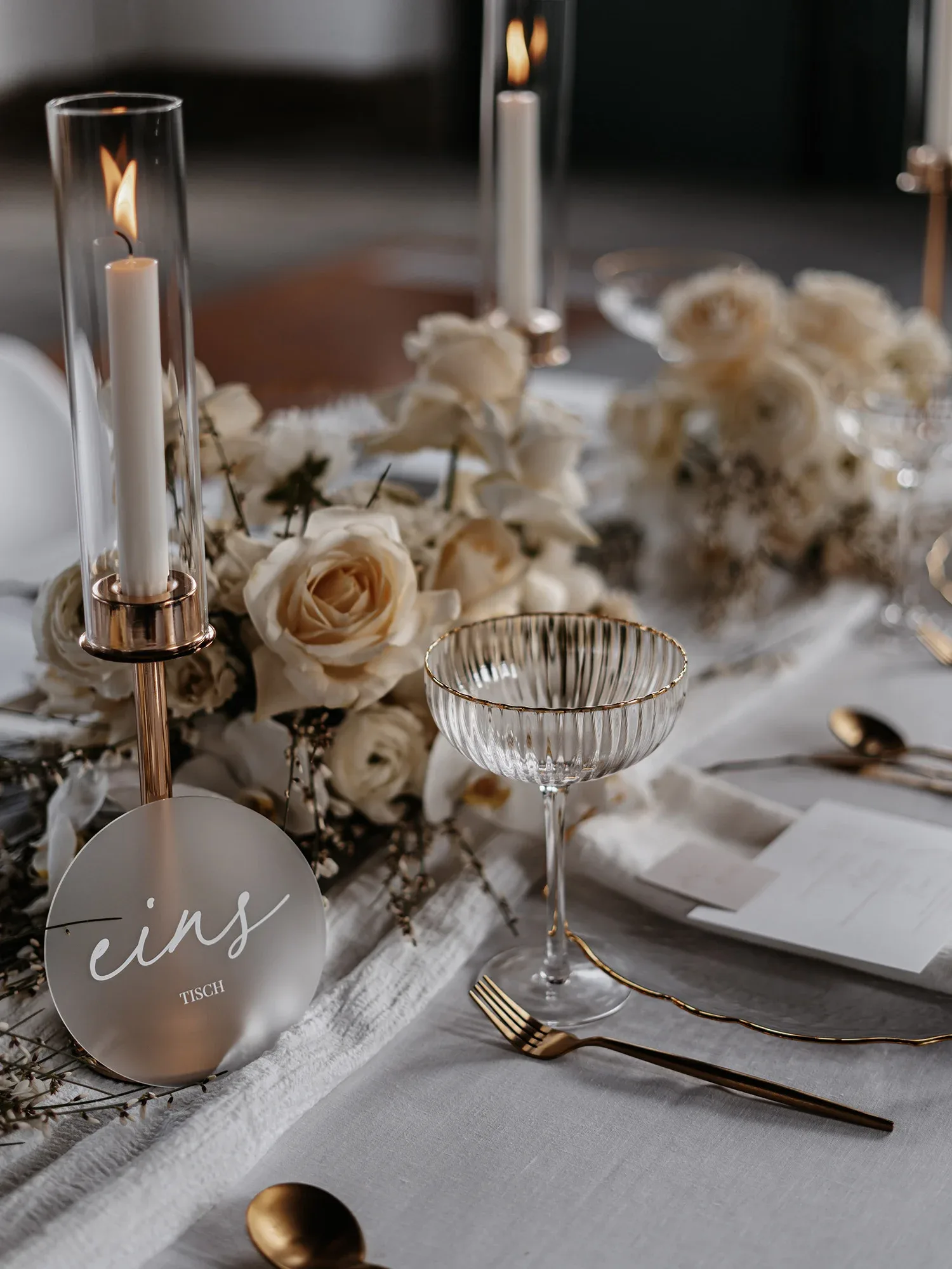 Elegante Hochzeitstischdekoration mit weißen Rosen, goldenen Bestecken und einer brennenden Kerze in einem Glaszylinder, daneben ein Tischschild mit der Aufschrift 'eins'.