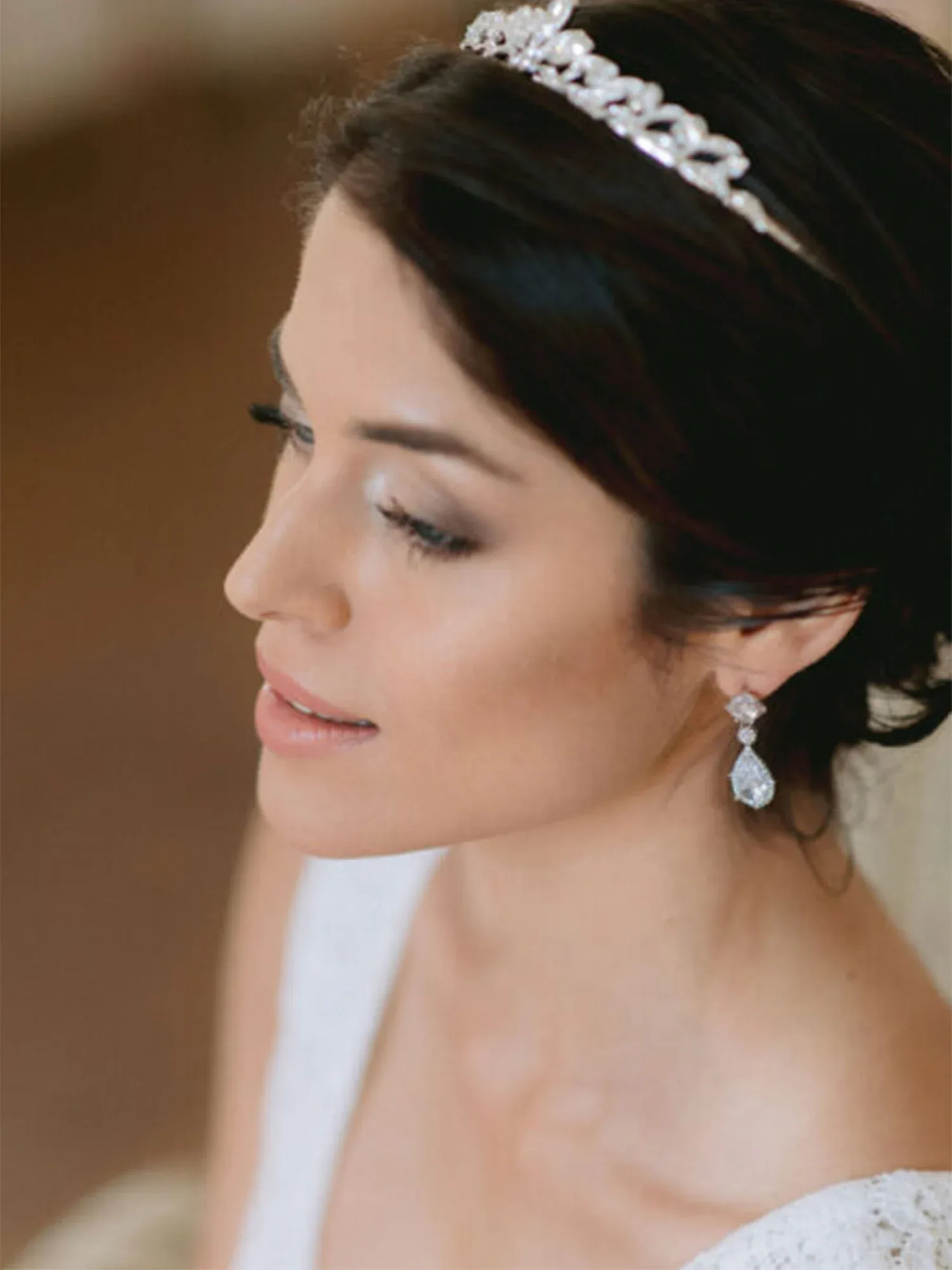 Eine Braut in einem weißen Spitzenkleid mit einer funkelnden Tiara und hängenden Diamantohrringen. Die Frisur ist elegant hochgesteckt.
