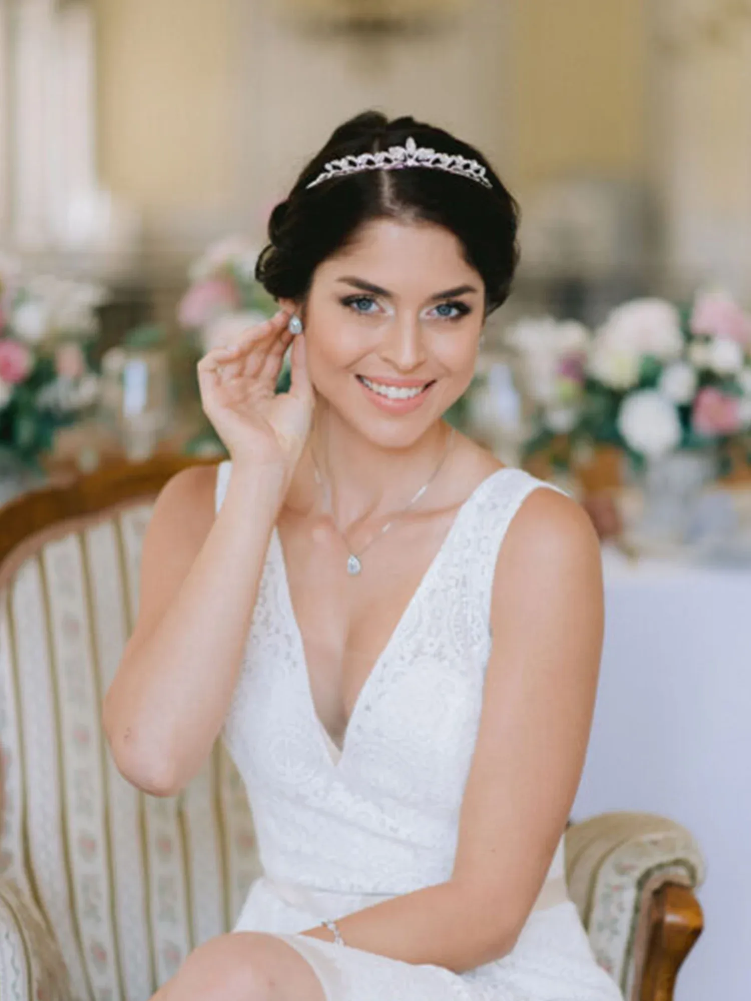 Eine Braut in einem weißen Spitzenkleid sitzt auf einem antiken Sessel und trägt eine funkelnde Tiara, lächelnd und elegant geschmückt.